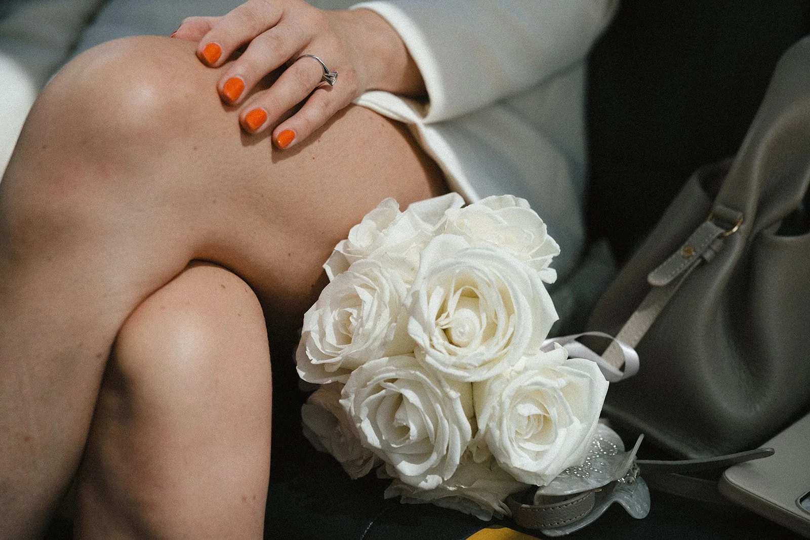 A woman with orange nail polish is sitting on a black chair, holding her knee with one hand, which has a ring on her finger. She is dressed in a white blazer. There is a bouquet of white roses resting on her lap, and a gray handbag is placed beside h