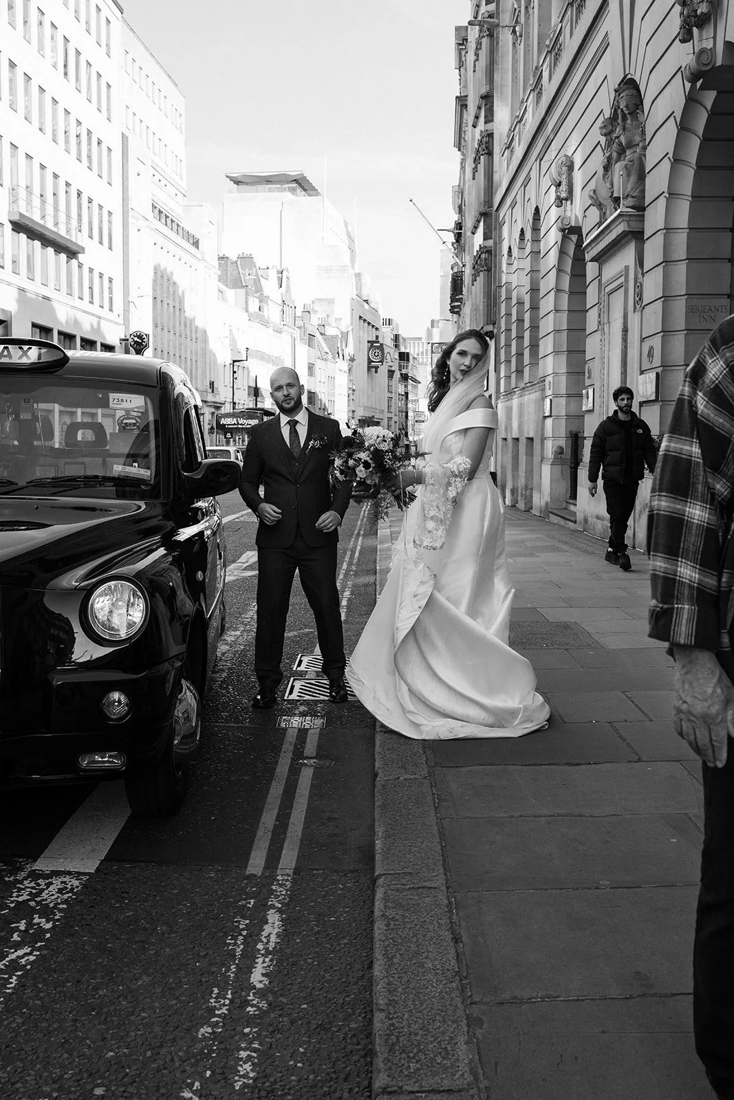A black-and-white photo of a bride and groom standing on a city street. The bride is holding a bouquet and wearing a wedding gown with lace sleeves, while the groom is in a suit. There are tall buildings and a taxi cab on the street, with a few pedes