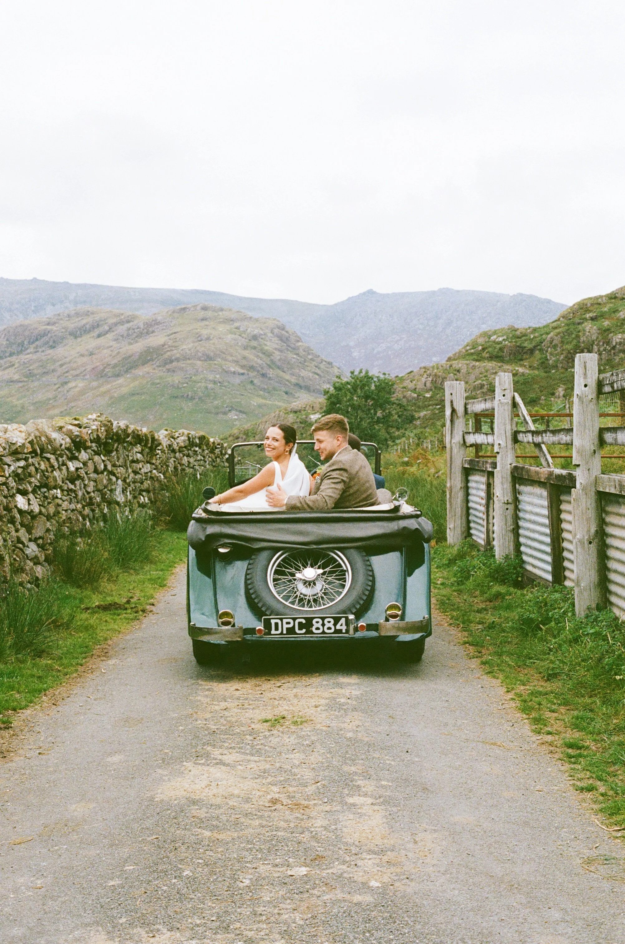A vintage car with a man and woman inside, driving through a scenic rural area with mountains and grassy fields in the background.
