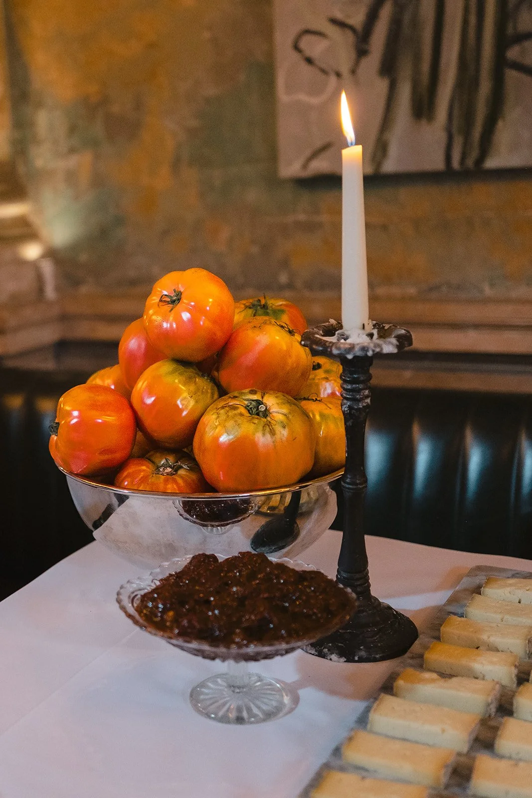 A table with a bowl of heirloom tomatoes, a lit candle in a candle holder, a glass dish of fig jam, and some small sandwiches.