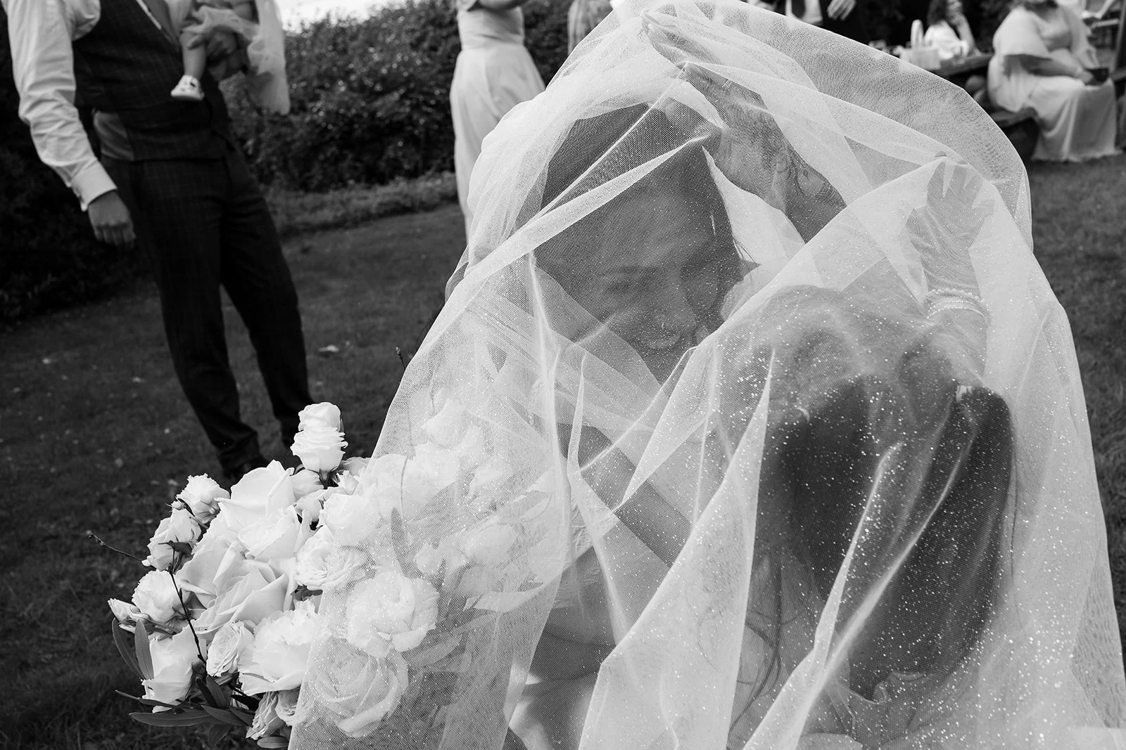 Bride in a veil sitting on the grass holding a bouquet of flowers, with guests in the background at an outdoor wedding.
