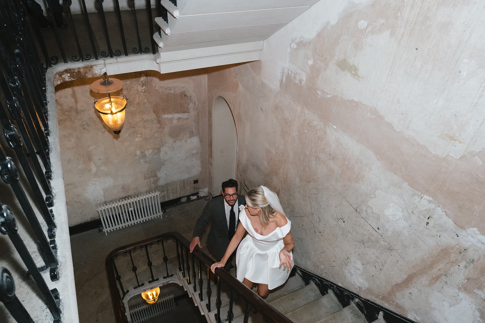 A bride in a white dress and a groom in a suit walking up a staircase in an old, rustic building with exposed brick walls and a vintage lamp hanging on the wall.