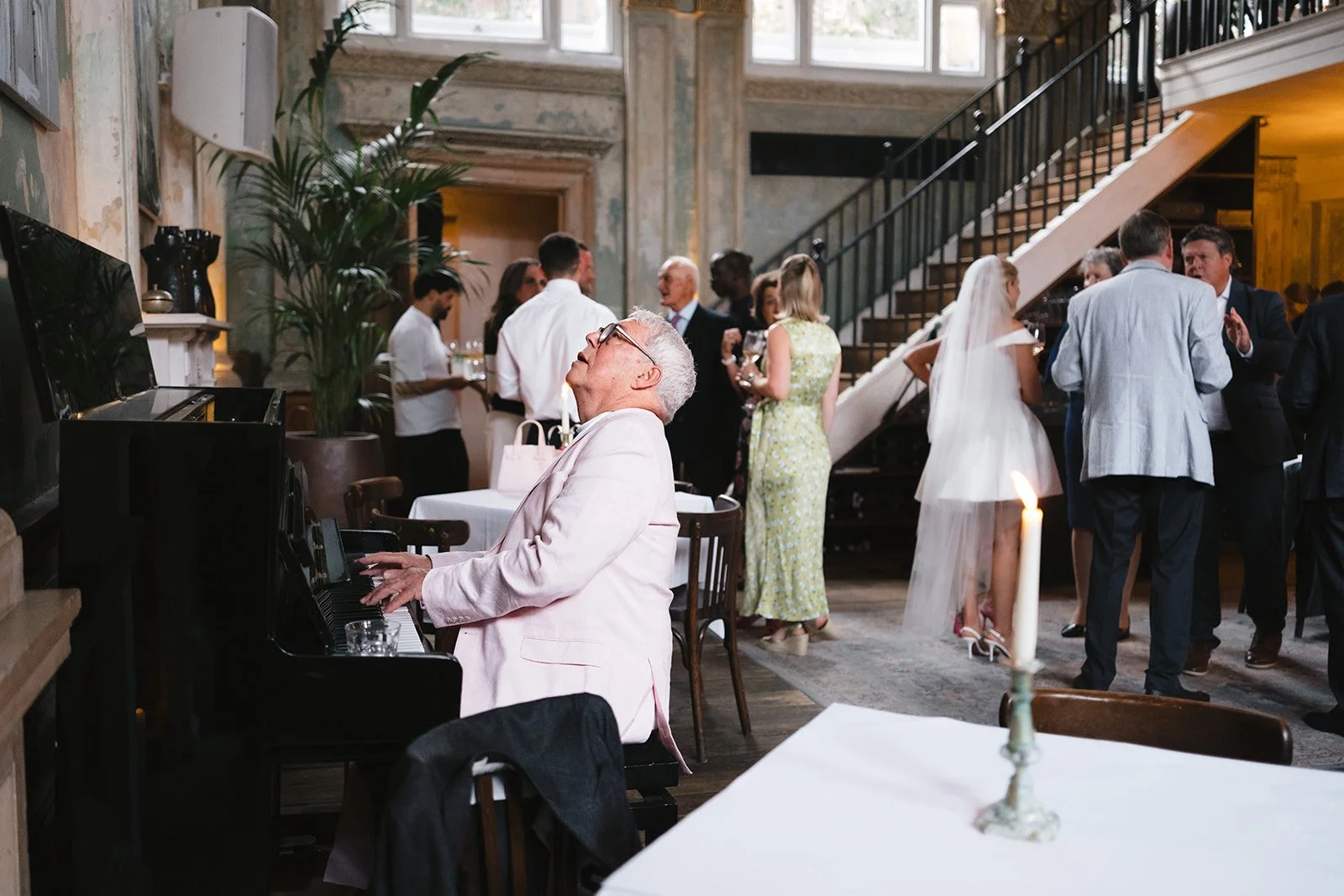 Older man in light pink suit playing a black piano at a wedding reception, with guests socializing in the background near a staircase.