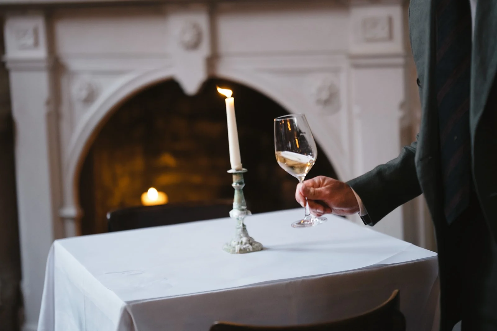 A man in a suit holding a glass of white wine near a table with a lit candle and a candle holder in a decorated room.