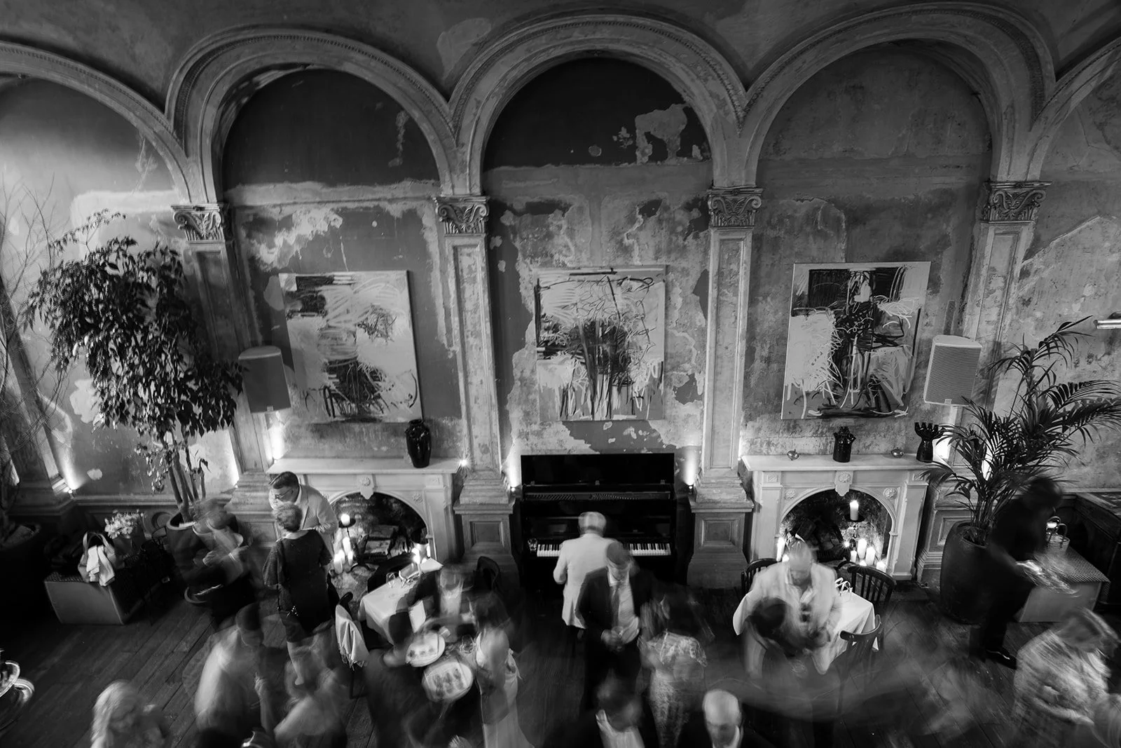 Black and white photo of a lively indoor gathering in an elegant room with high arched ceilings, large paintings, and a fireplace. People are standing, talking, and moving around, with a piano against the wall.