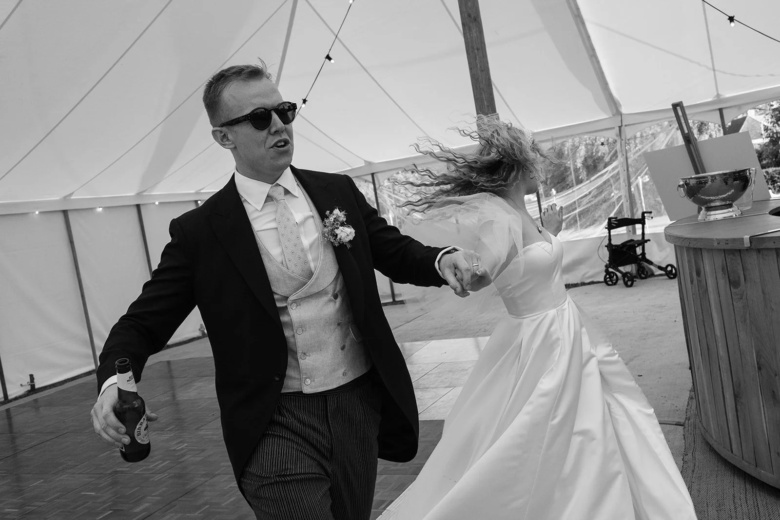 A black and white photo of a wedding celebration, showing a man in a tuxedo and sunglasses holding a beer bottle, dancing with a bride in a wedding dress inside a large tent decorated with string lights.