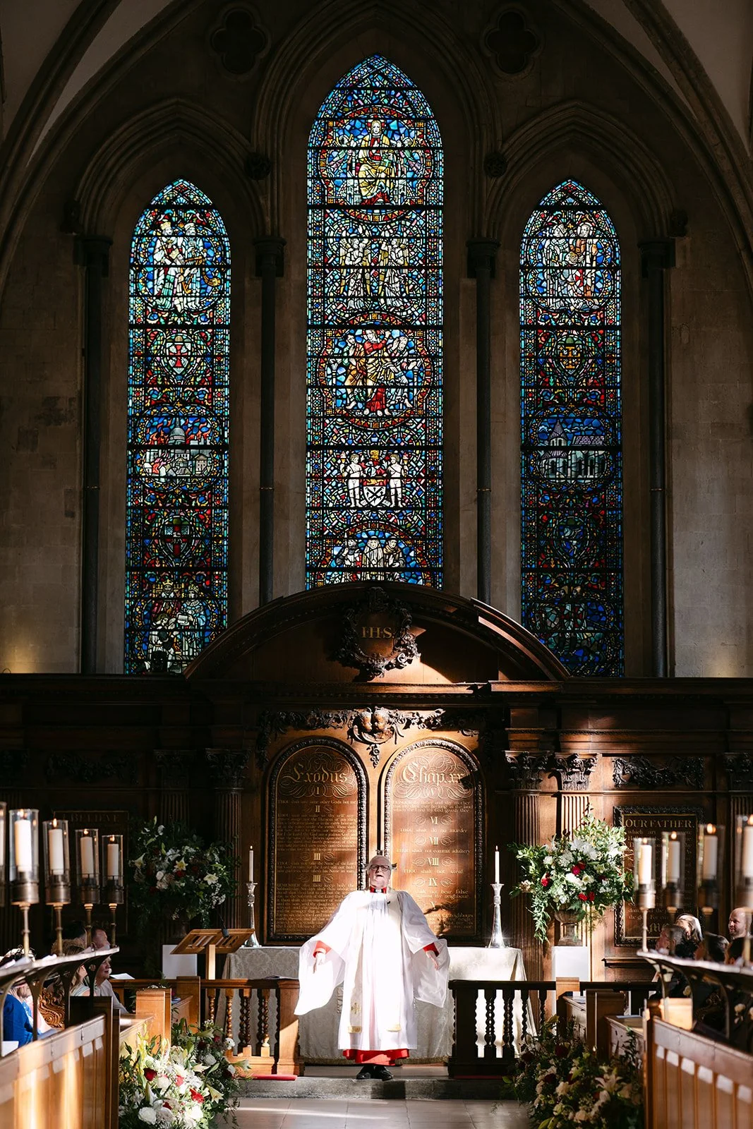 A priest in white robes standing at the altar of a church, with three tall stained glass windows behind him, decorated with religious images and symbols. Flowers and candles are on either side of him.