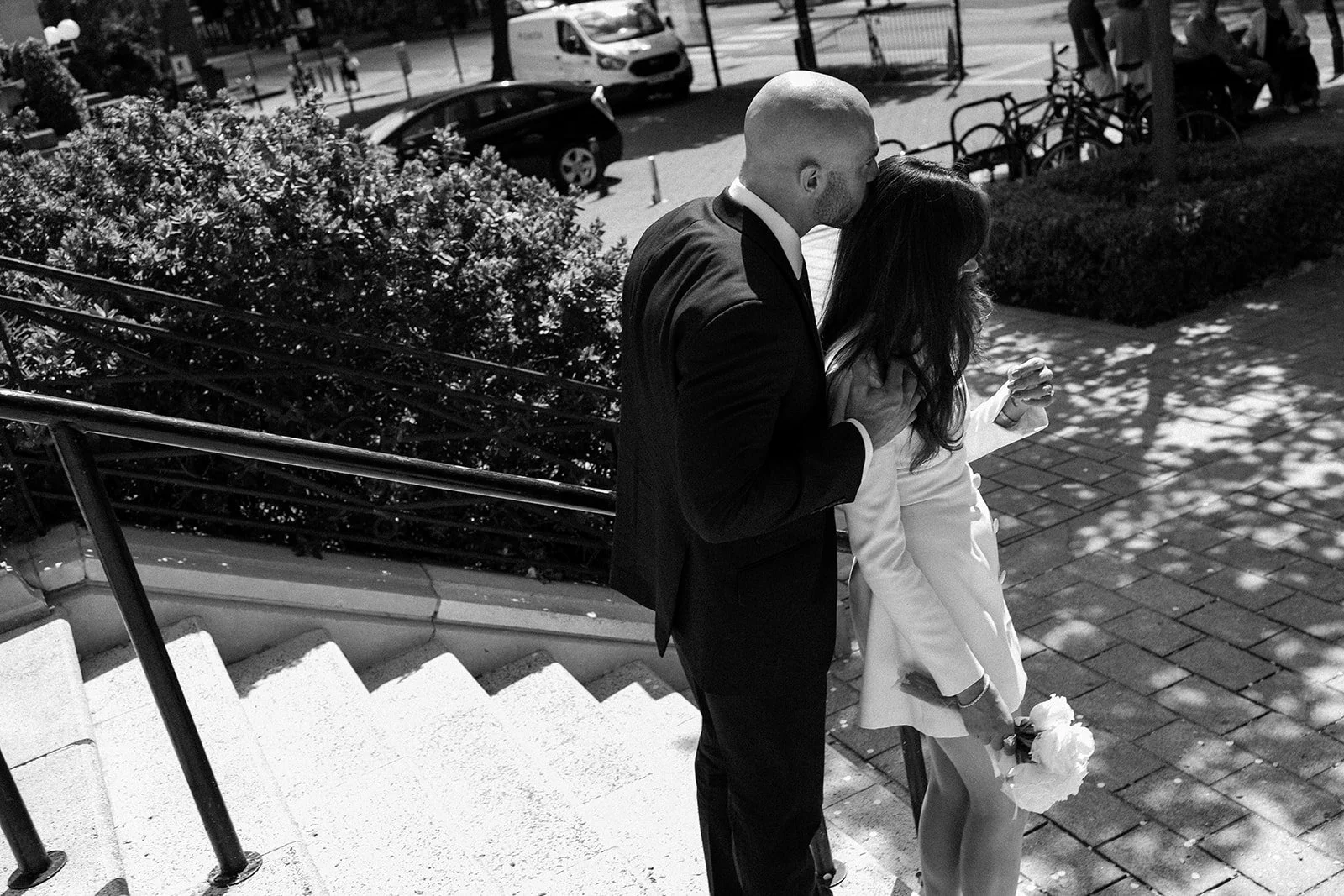 A couple, dressed formally, sharing a kiss on a city sidewalk, with the woman holding flowers.