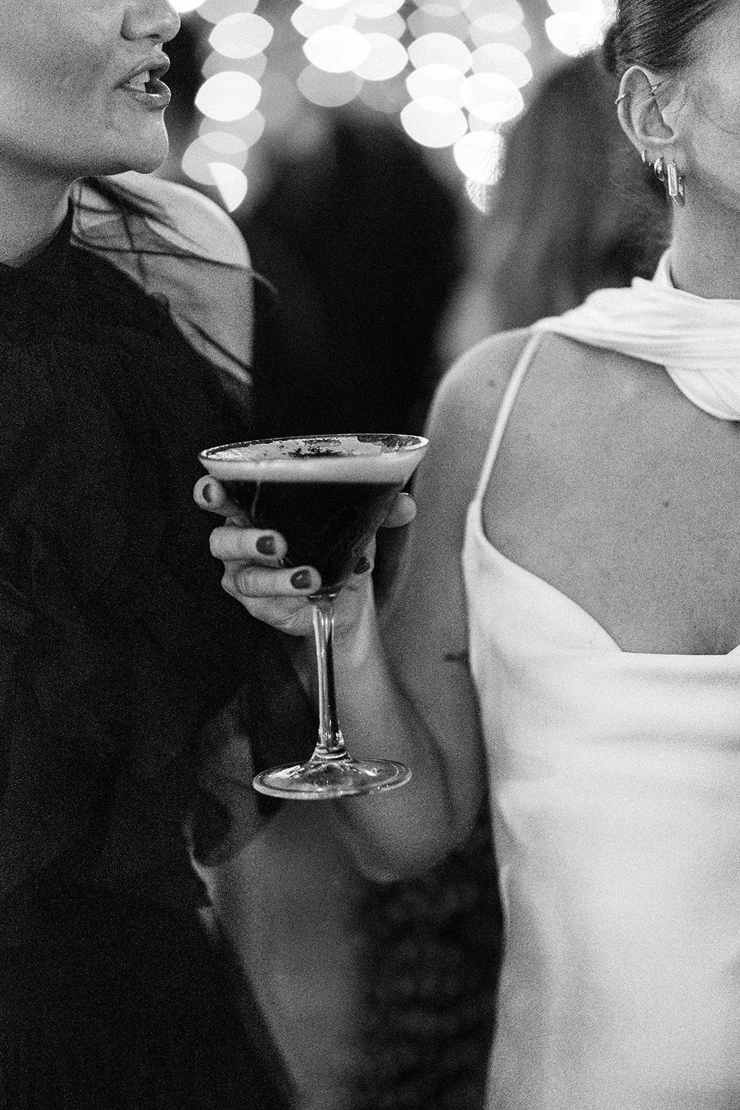 Close-up of two women at a social event, one holding a cocktail glass and the other wearing earrings, with blurred lights in the background.