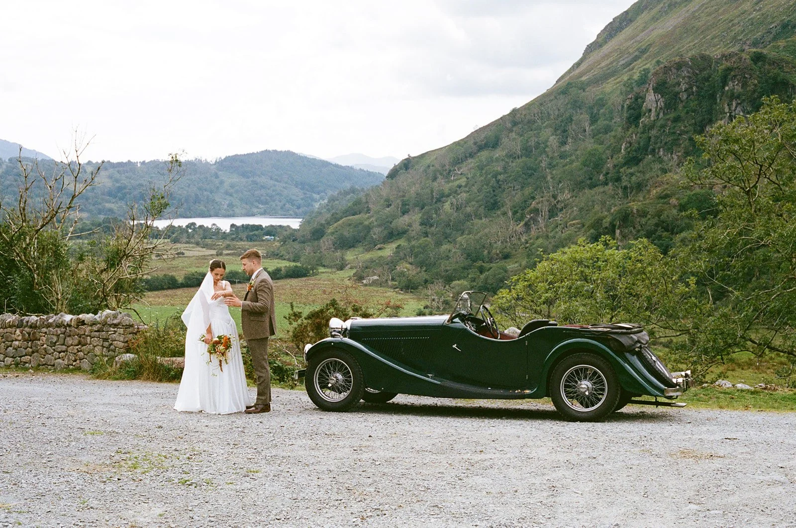 A bride and groom stand outdoors near a vintage black convertible car, with green hills and a lake in the background.