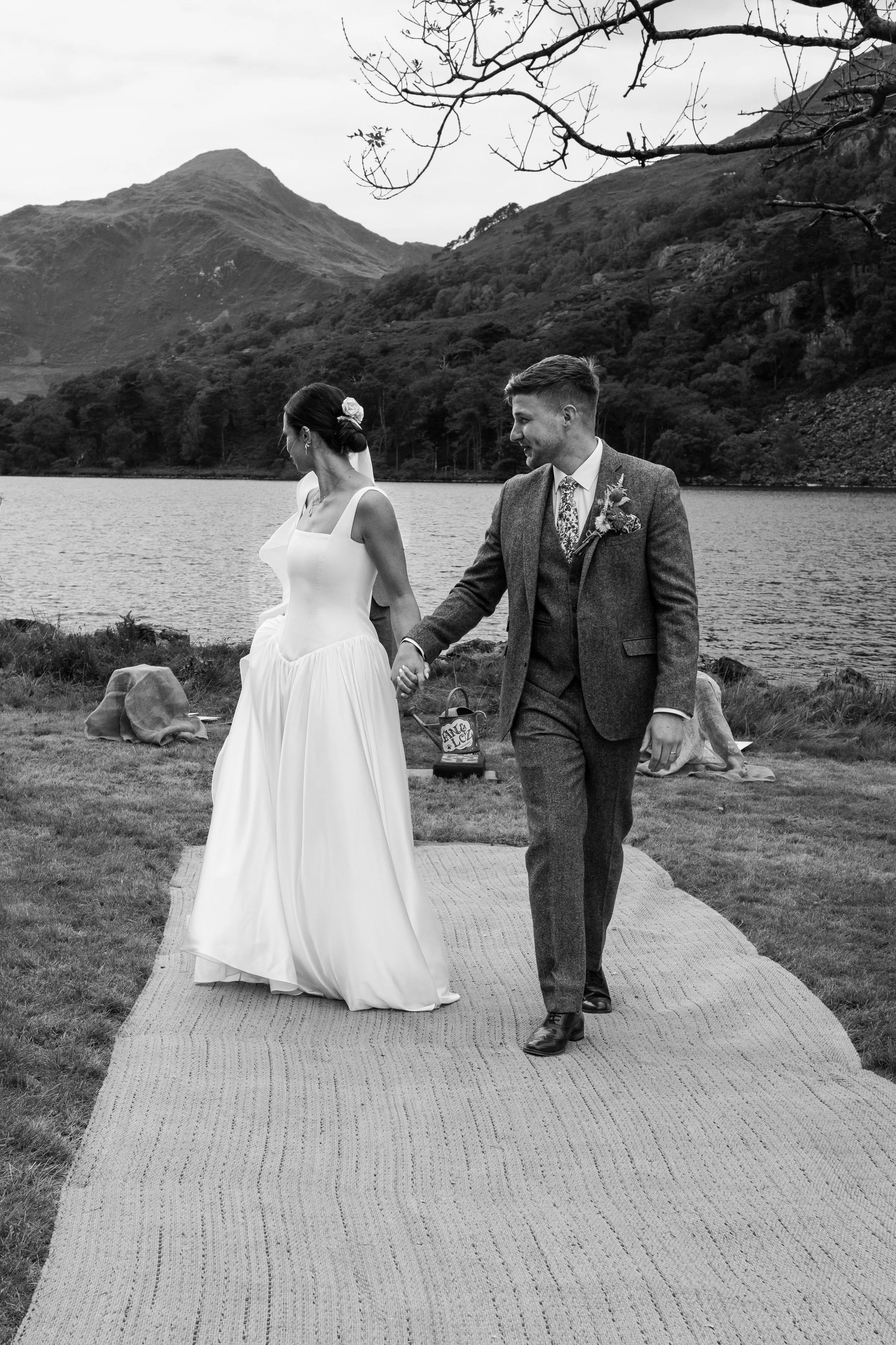 A black and white photo of a bride and groom holding hands during an outdoor wedding ceremony near a lake with hills in the background.