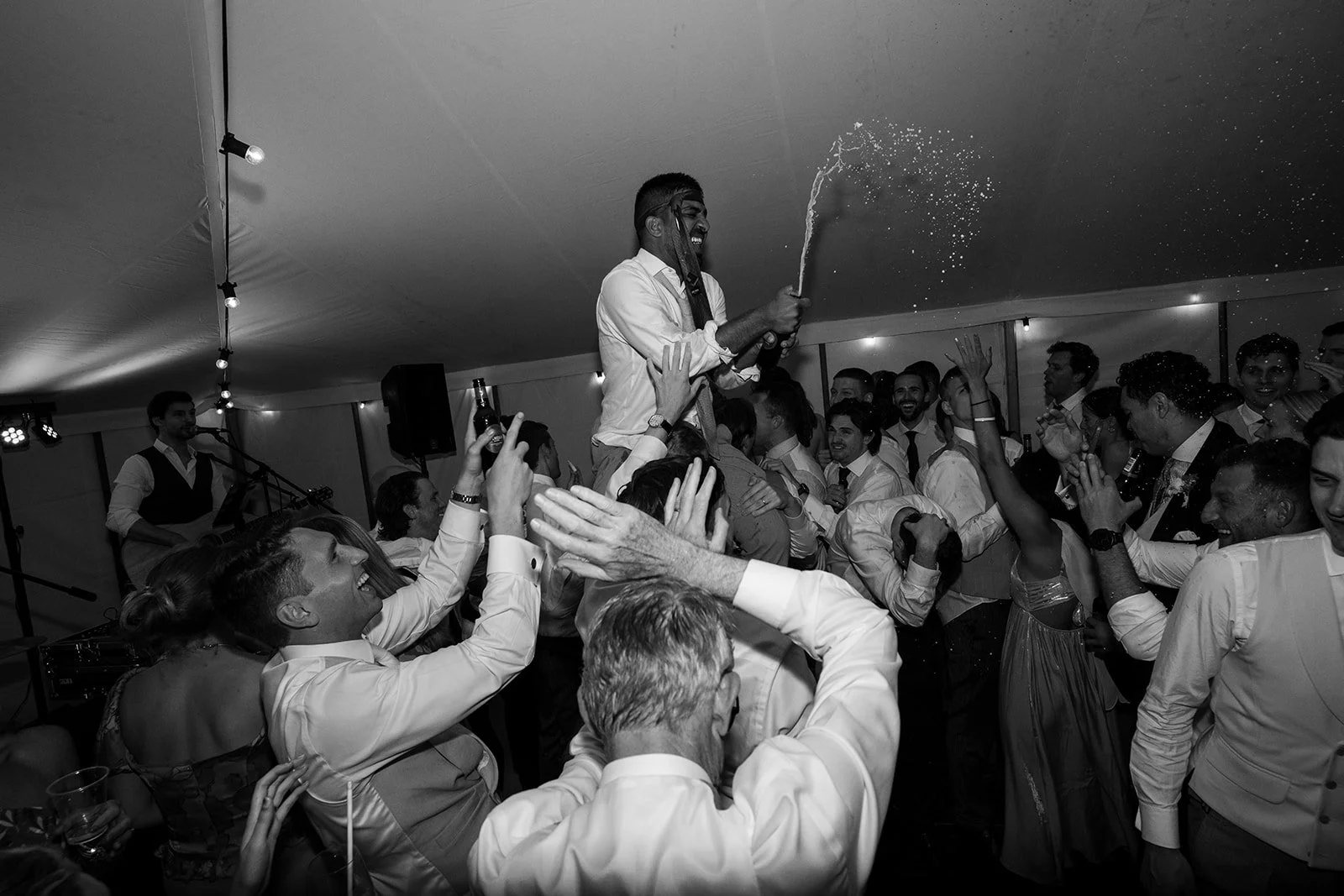People celebrating at a lively party, with one person being lifted on others' shoulders, spraying a drink into the air. Everyone appears happy and engaged, with some holding drinks and dancing.