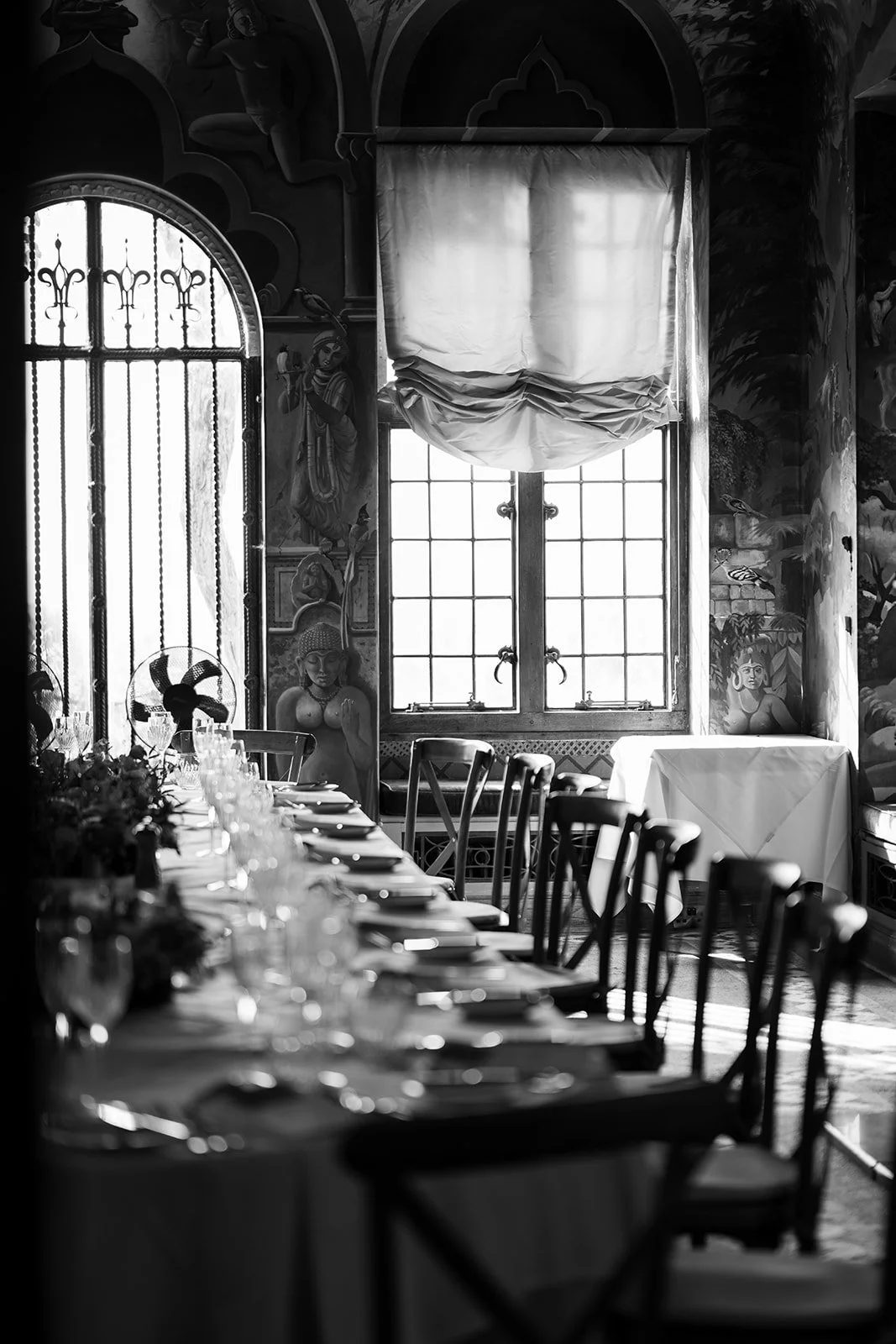 A black and white photo of a long dining table covered with a tablecloth, set with glasses, plates, and silverware, inside a decorated room with ornate paintings on the walls, large windows with curtains, and wooden chairs around the table.
