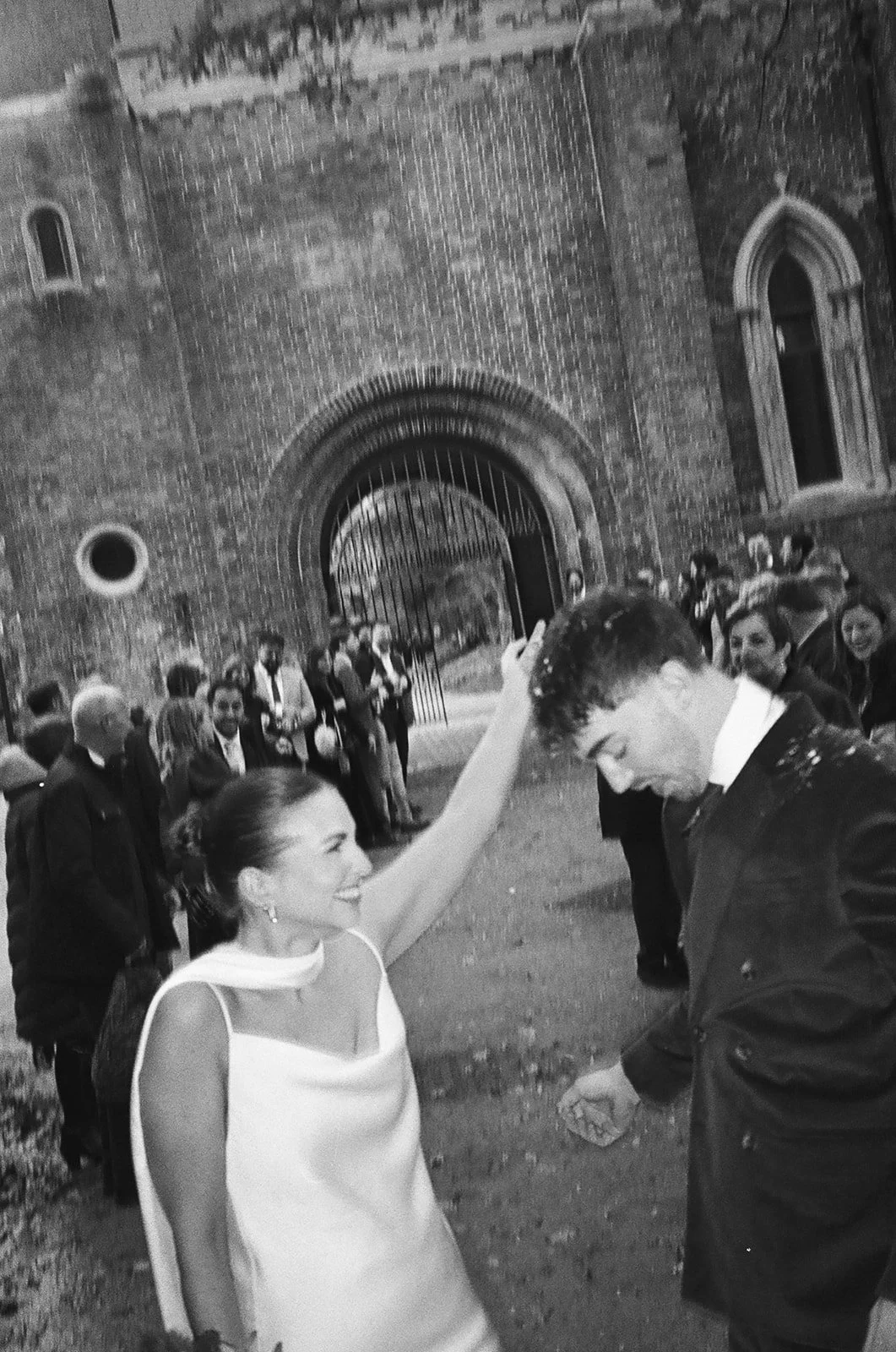 A woman in a sleeveless white dress is smiling and touching the forehead of a young man in a dark suit, outside a historic brick building with an arched gate, surrounded by a group of people.