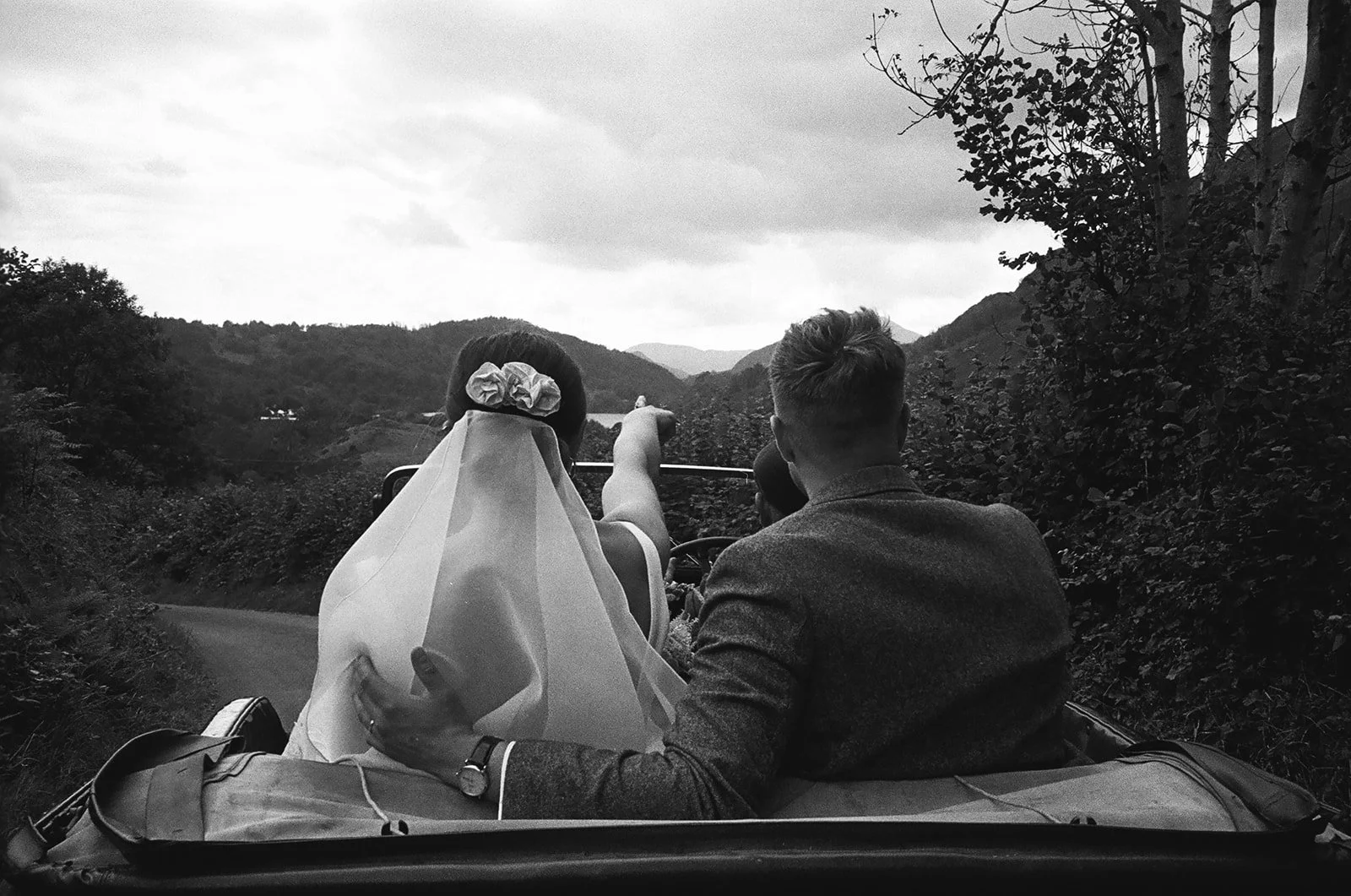 A black and white photo of a couple riding in a vintage car through a scenic hilly landscape, with the woman wearing a veil and flowers in her hair, and a man sitting beside her.