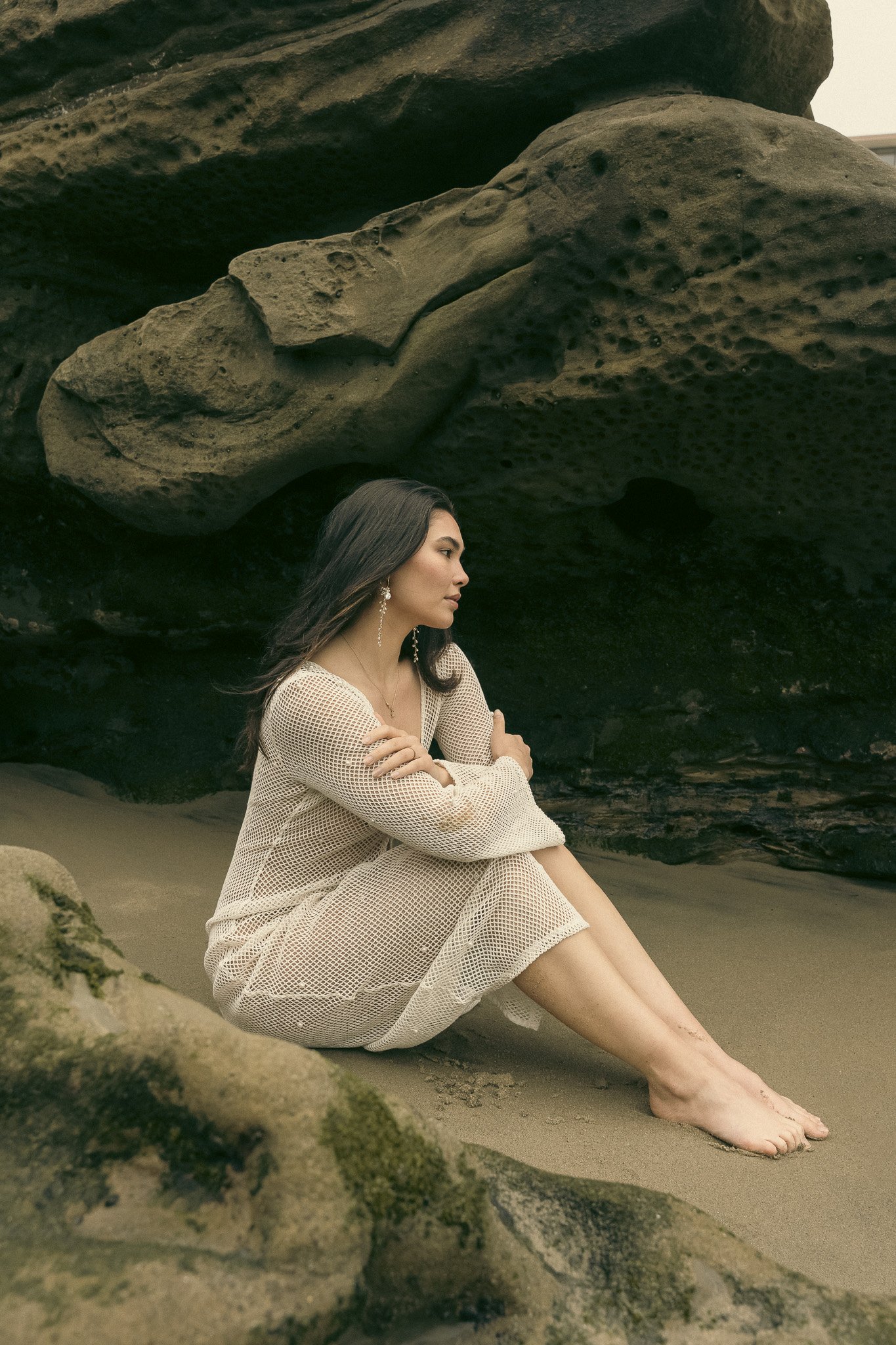 Woman in a white mesh dress sitting on a sandy beach near rocky formations.