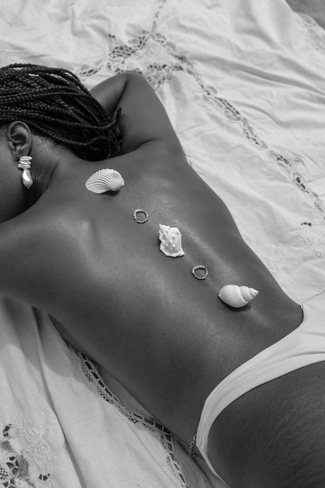 Black and white photo of a person lying on a bed with shells and rings arranged on their back.