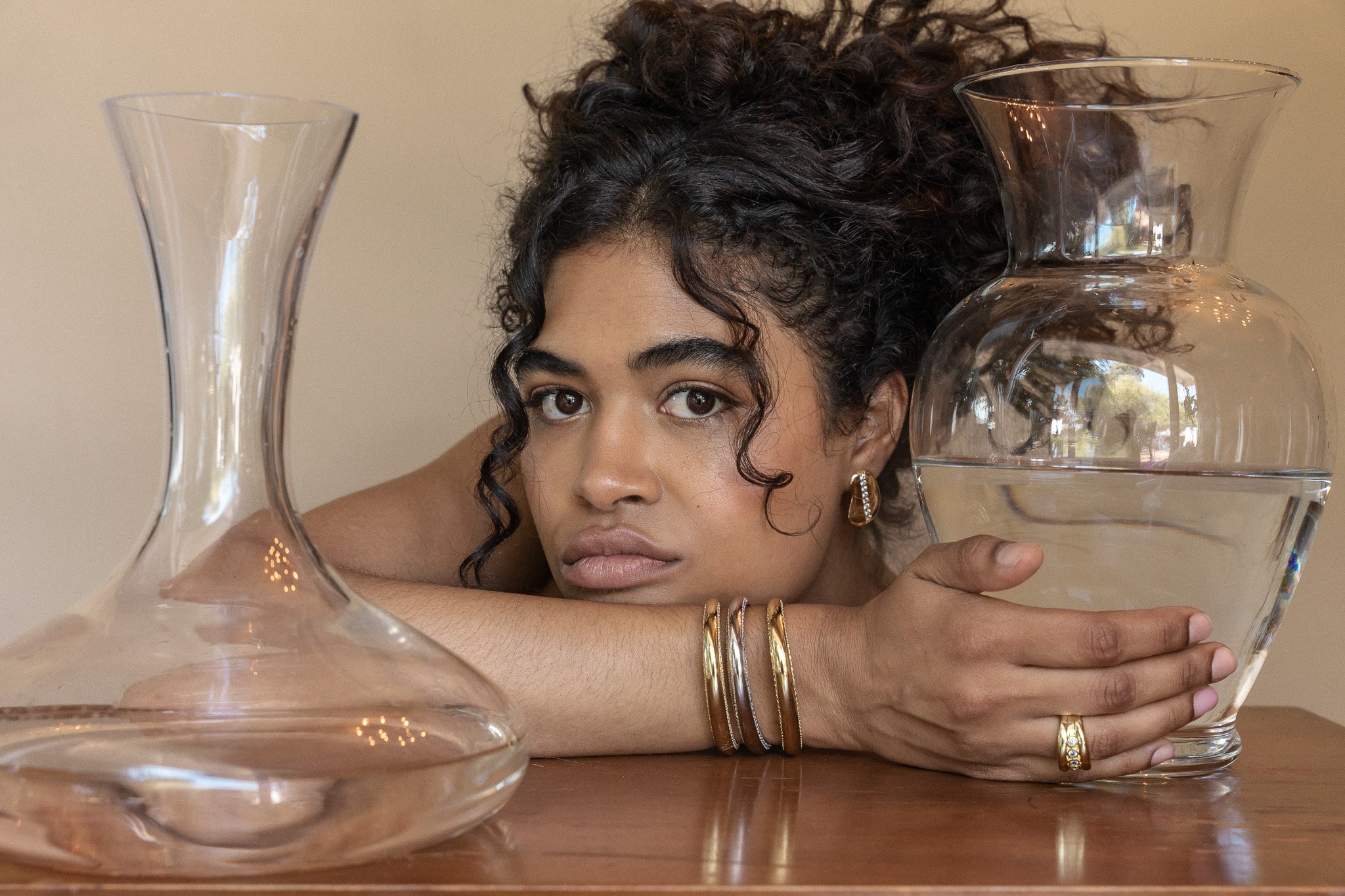 Woman with curly hair, head resting on a table, holding a glass vase half-filled with water, next to an empty glass carafe.