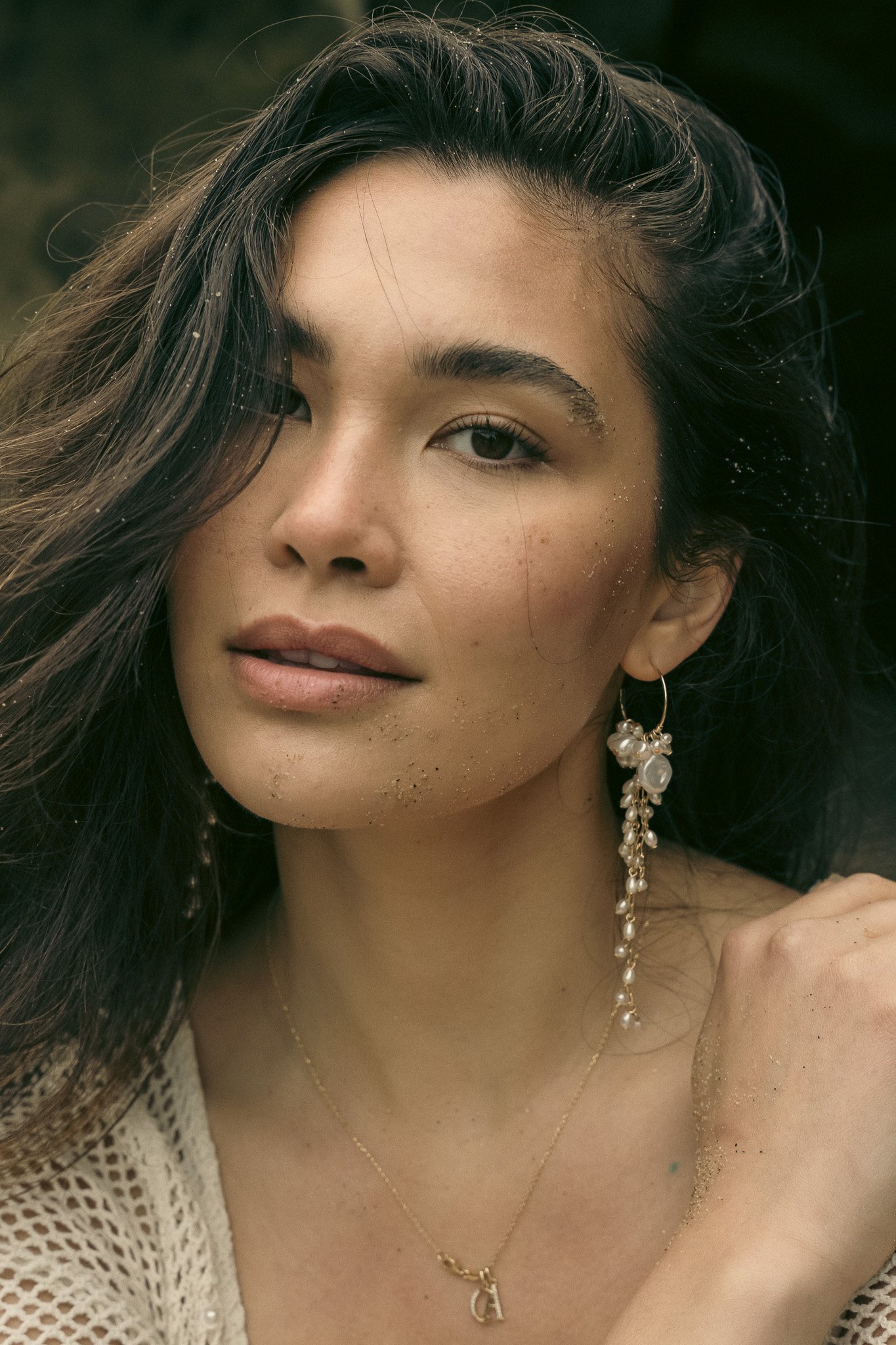 Close-up of a woman with light sand on her face, wearing a long pearl earring and a thin necklace.