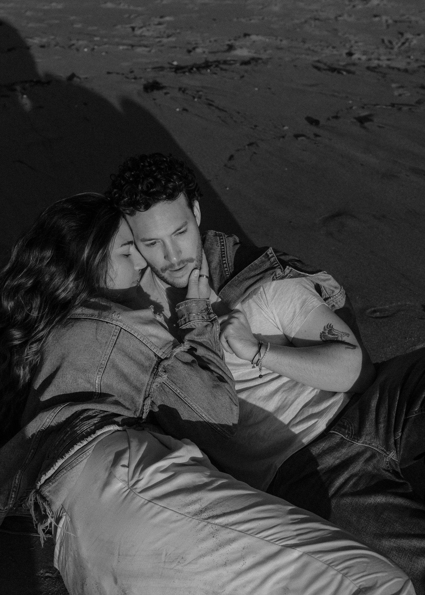 A black and white photo of a couple cuddling on sand. The man is lying down wearing a T-shirt and jeans, while the woman is resting her head on his shoulder in a denim jacket. They look relaxed and affectionate.