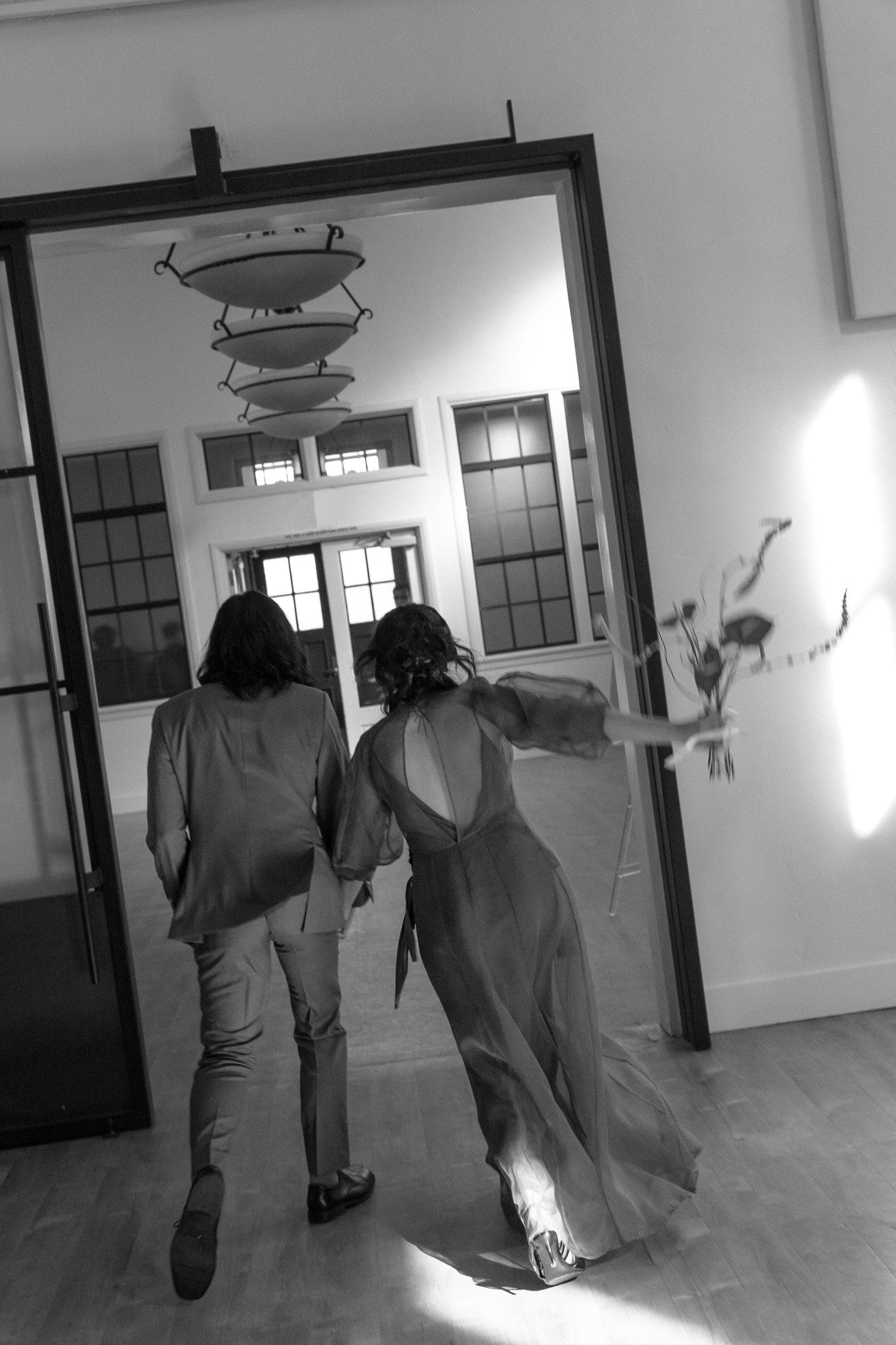 Black and white photo of a couple dressed formally, walking hand in hand towards a brightly lit room, with the woman holding a bouquet.