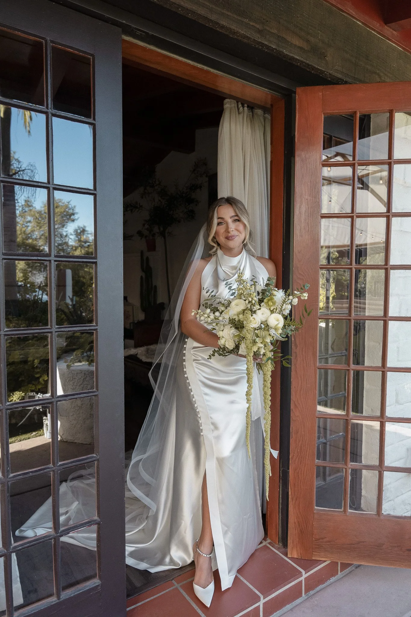 Bride in a white satin wedding dress with a train, holding a bouquet of white flowers, standing in a doorway.