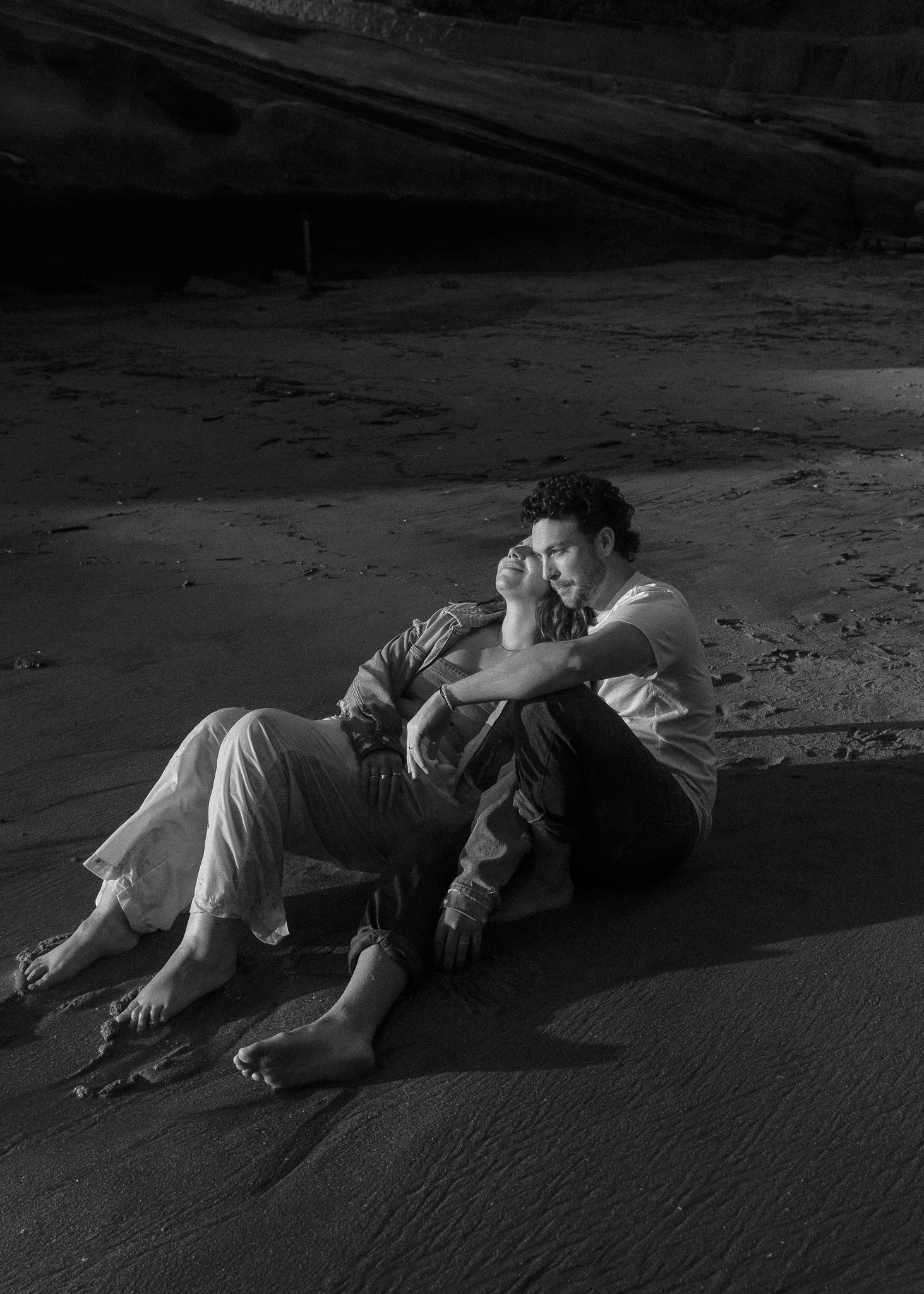 Two people sitting on a beach at night, leaning against each other, with one person smiling and the other looking contemplative.