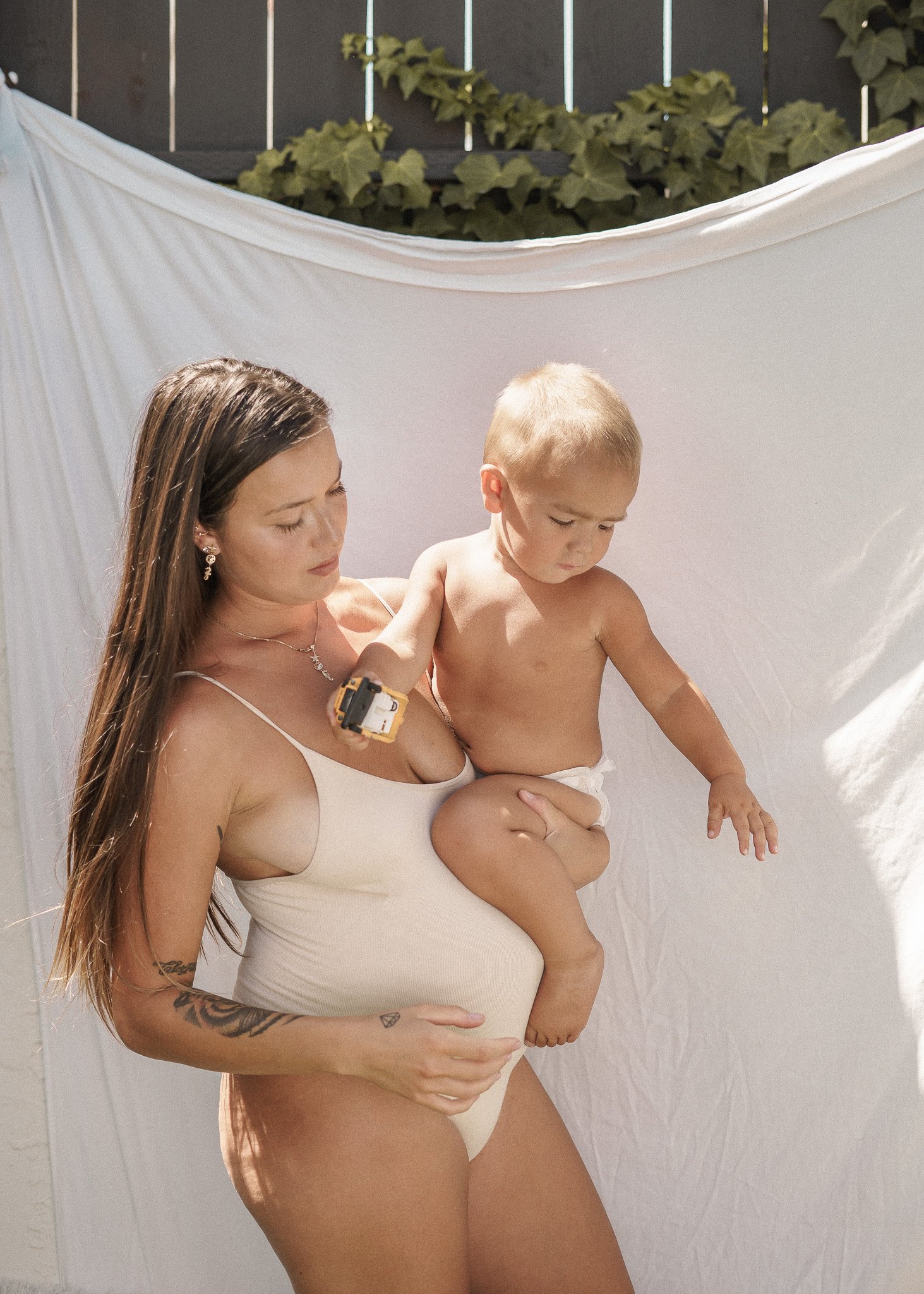 Pregnant woman in swimsuit holding toddler against white backdrop outdoors