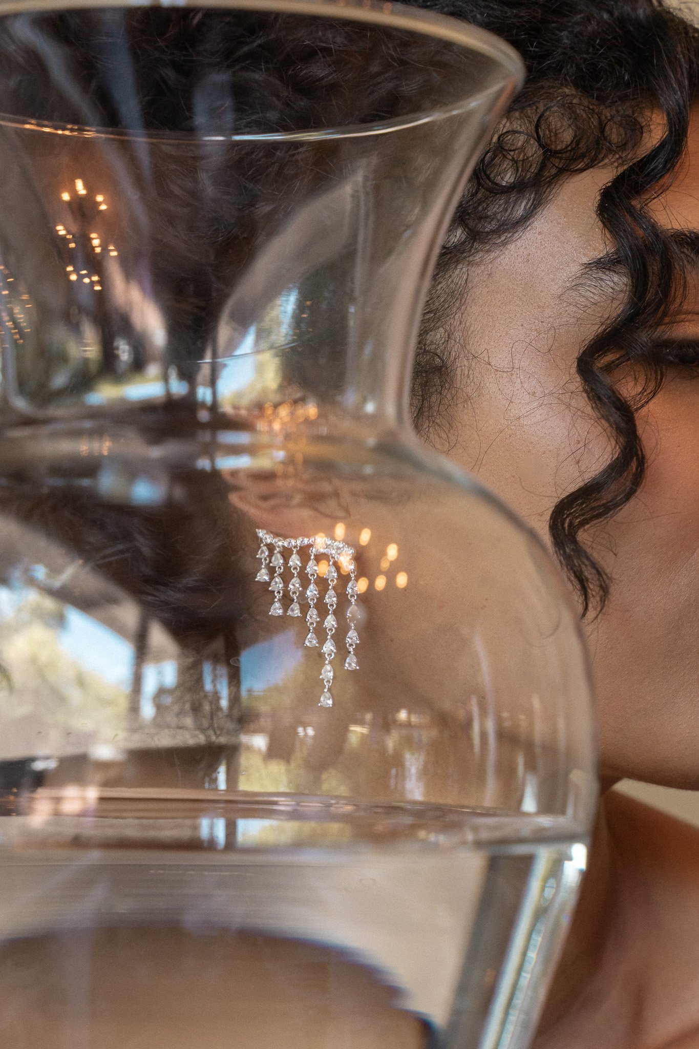 Close-up of a woman wearing sparkling earrings, partially obscured by a reflective glass vase filled with water, with elegant lighting reflections.