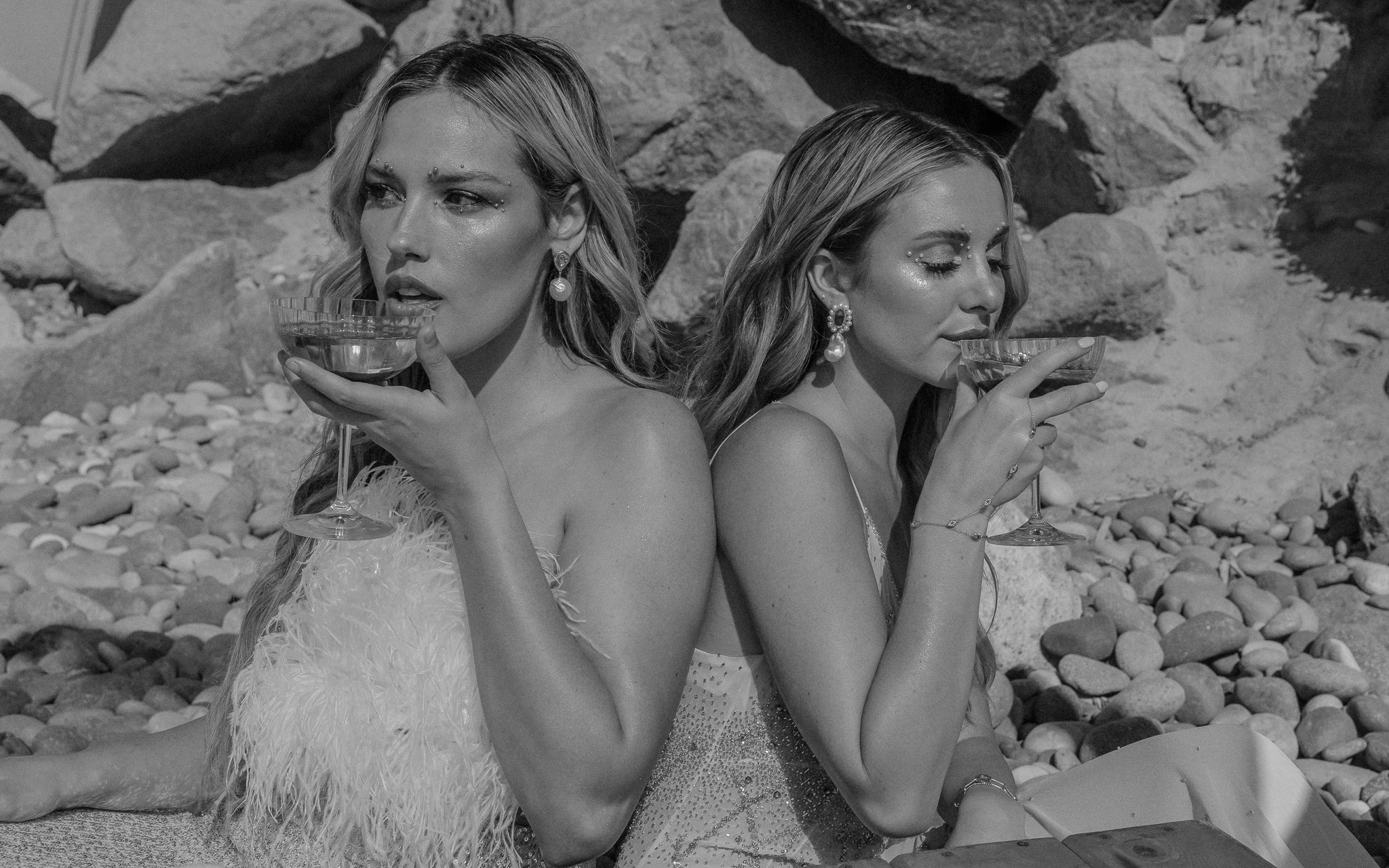 Two women sitting back to back on a pebble beach, each holding a coupe glass with a drink, wearing elegant clothing and pearl earrings, black and white photo.