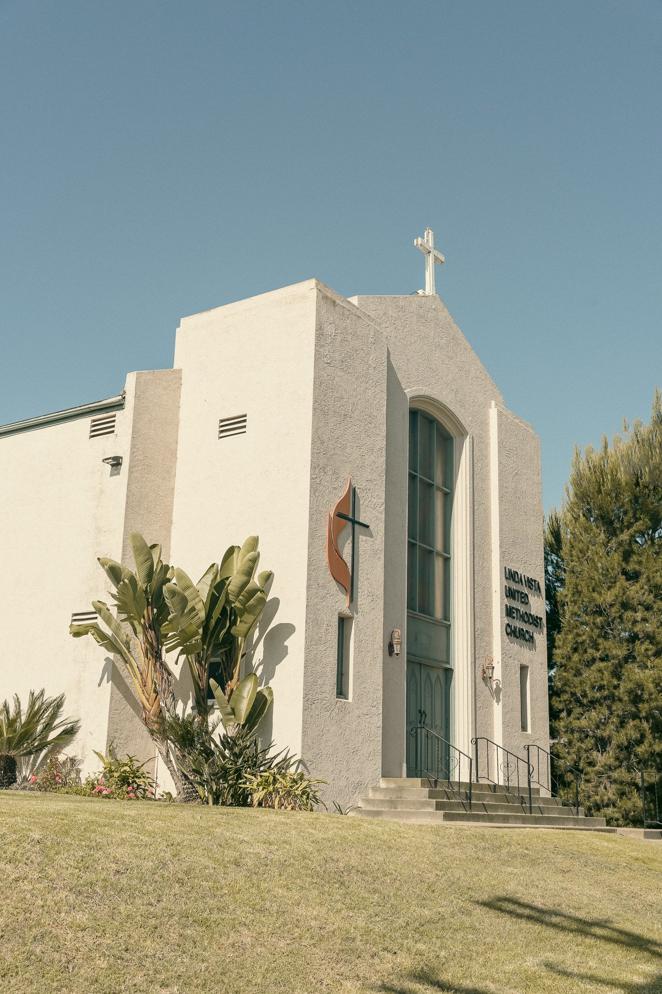 A white church building with a cross on top, featuring the sign "Linda Vista United Methodist Church" on the front. The entrance is marked by a large decorative window and stairs, with greenery surrounding the base.