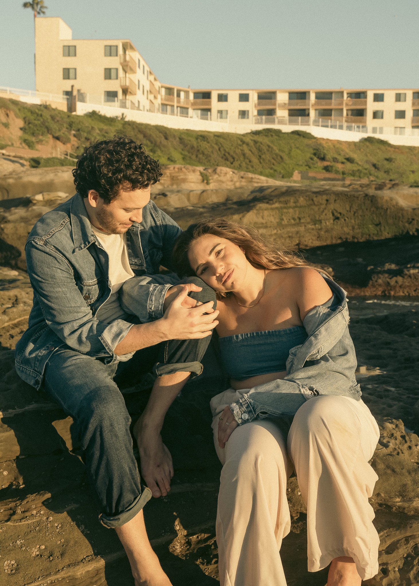 A couple sitting on rocks near a beach, with a building in the background, wearing casual denim and light-colored clothes.
