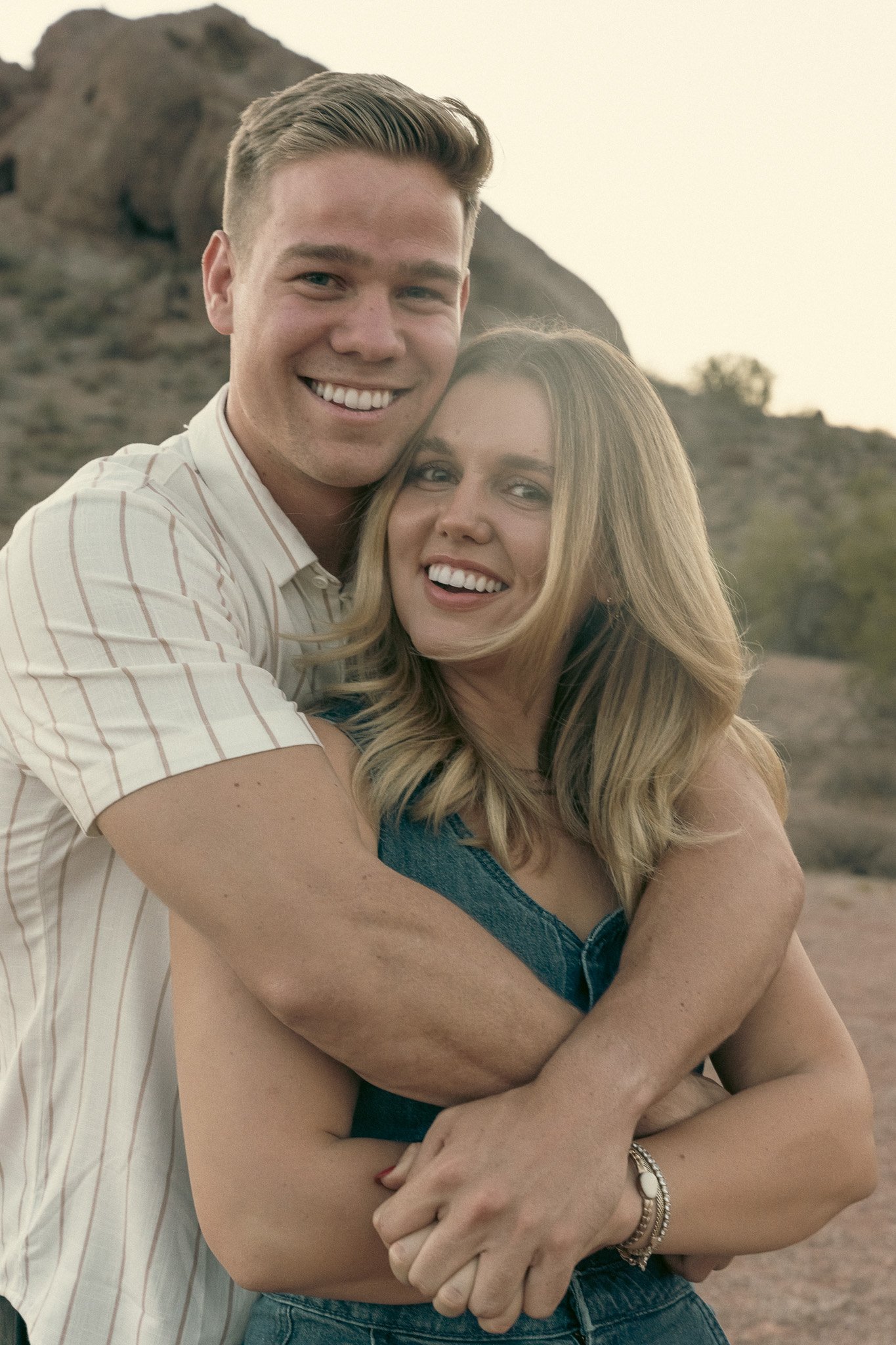 A couple smiling and embracing in a desert landscape with a mountain in the background.