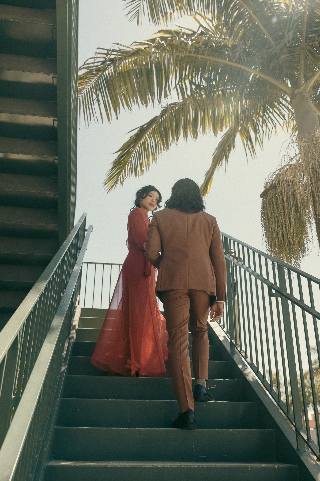 A woman in a red dress and a man in a suit walking up outdoor stairs, with palm trees in the background.