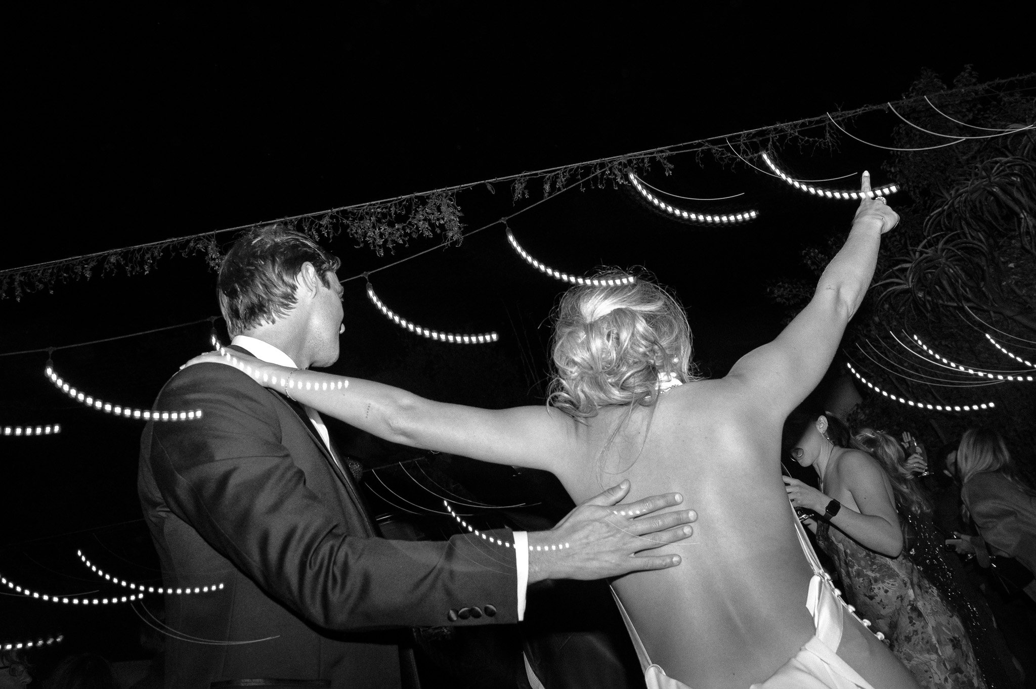 Black and white photo of a couple dancing at a party. The man is in a suit, and the woman is wearing a backless dress. String lights are visible above them, creating a festive atmosphere.