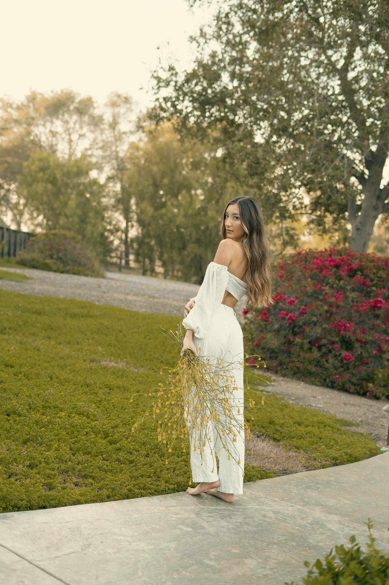 Woman in white dress holding yellow flowers in a garden, surrounded by greenery and trees.