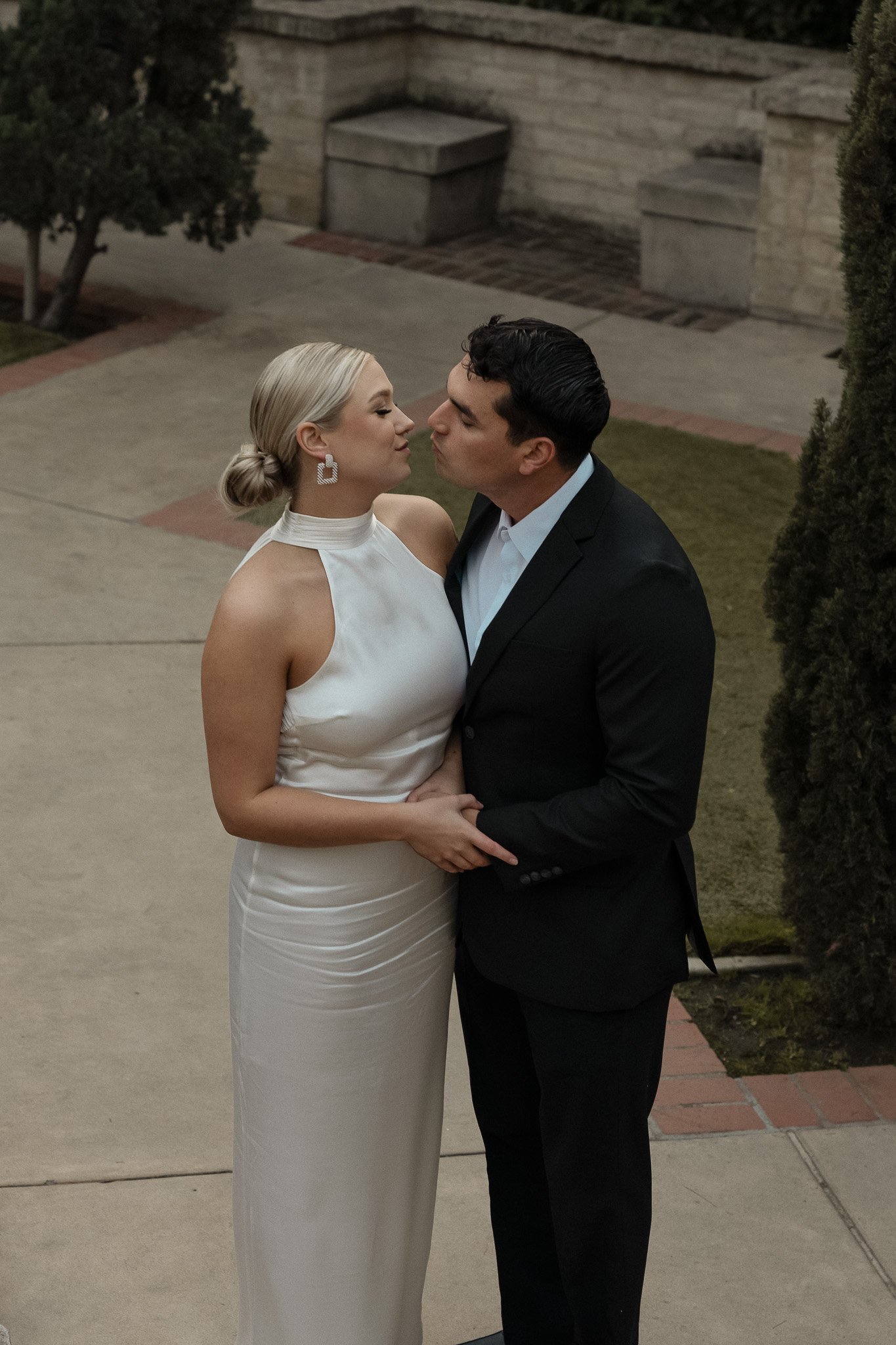 A couple embracing in an outdoor setting, the woman in a white dress and the man in a black suit, appearing to kiss.
