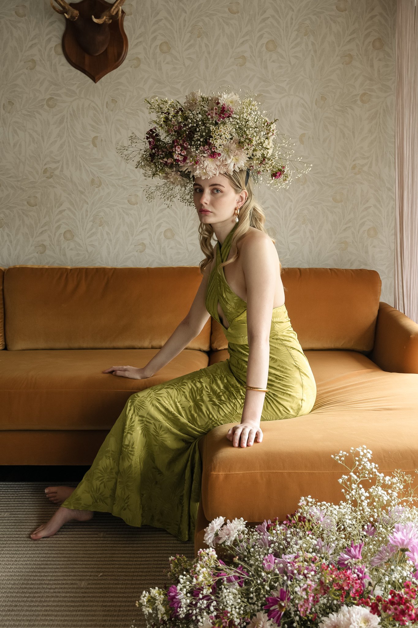 Woman in green dress with floral headpiece sitting on mustard couch with flowers.
