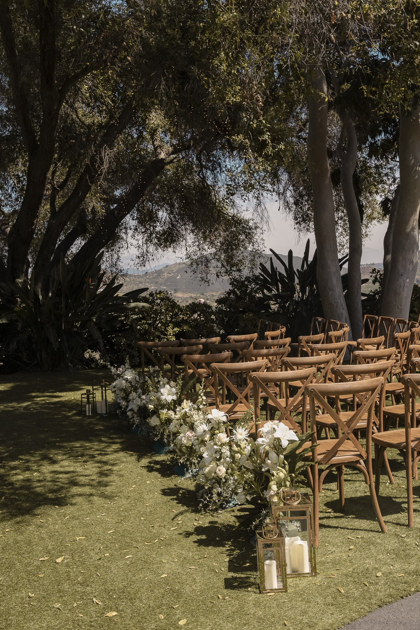 Outdoor wedding ceremony setup with wooden chairs arranged in rows, flower arrangements along the aisle, lanterns with candles, and large trees providing natural shade.