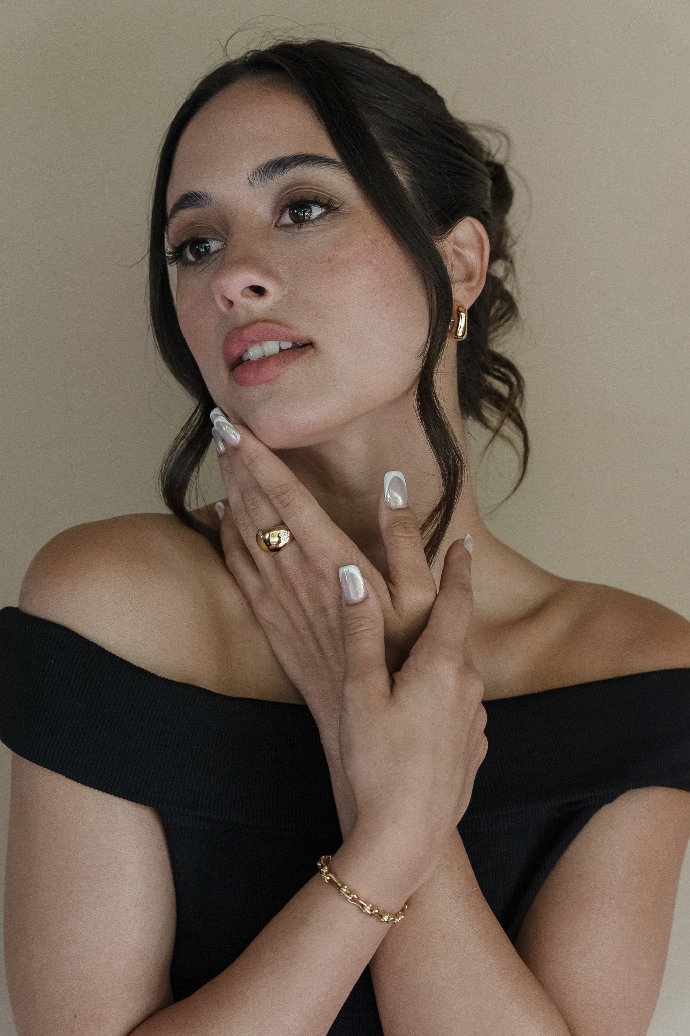 Woman with long dark hair wearing earrings, a gold ring, and a gold bracelet, posing with hands near face in black off-shoulder top.