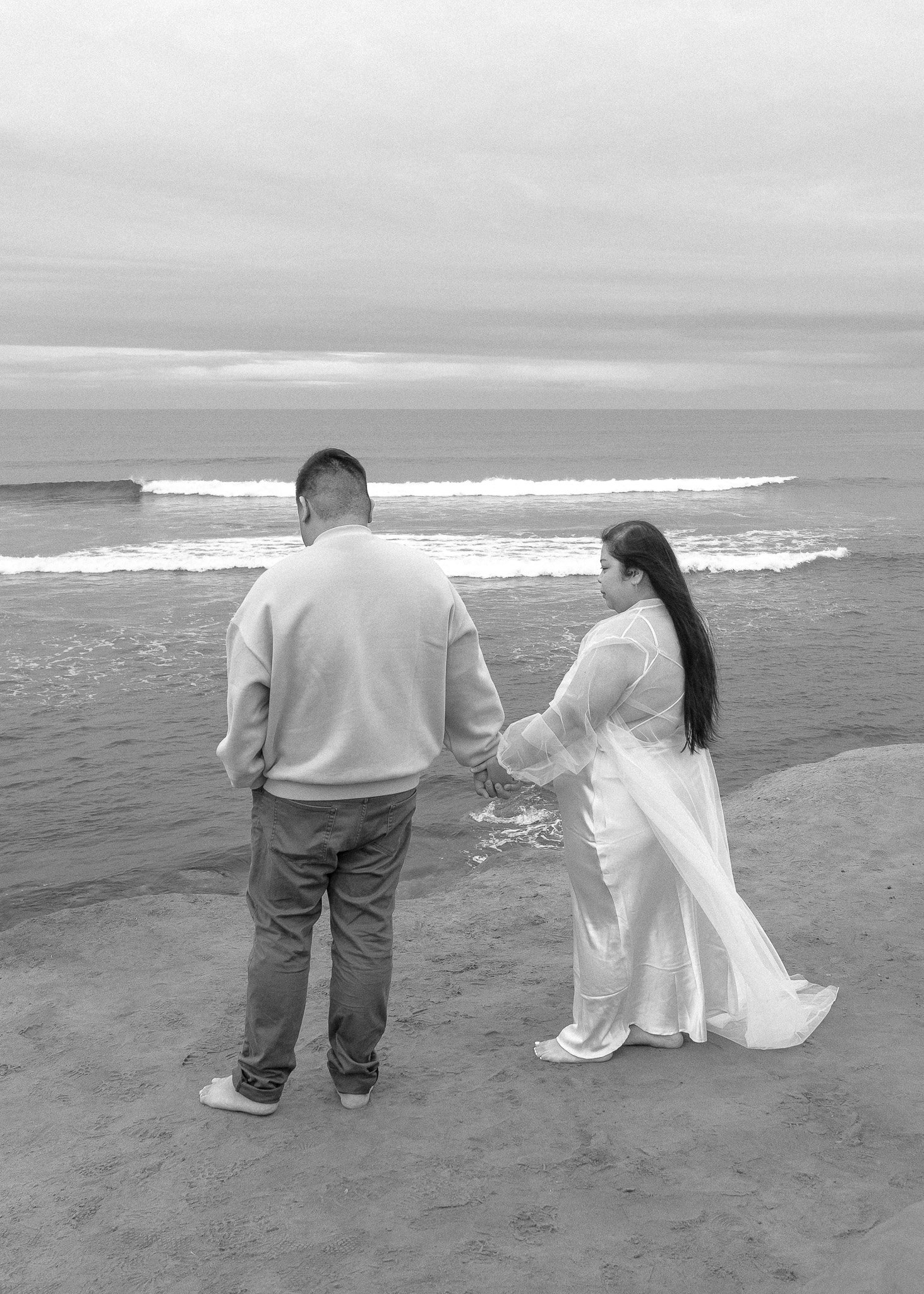 A black-and-white photo of a couple holding hands, standing on a sandy beach facing the ocean. The woman is wearing a long white dress, while the man is in casual clothing. The waves are gently crashing in the background.