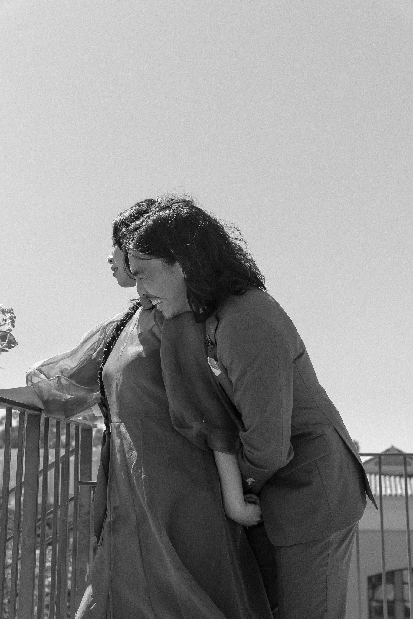 Two people standing together, one wearing a dress and the other in a suit, smiling and leaning over a railing, outdoors with a clear sky.
