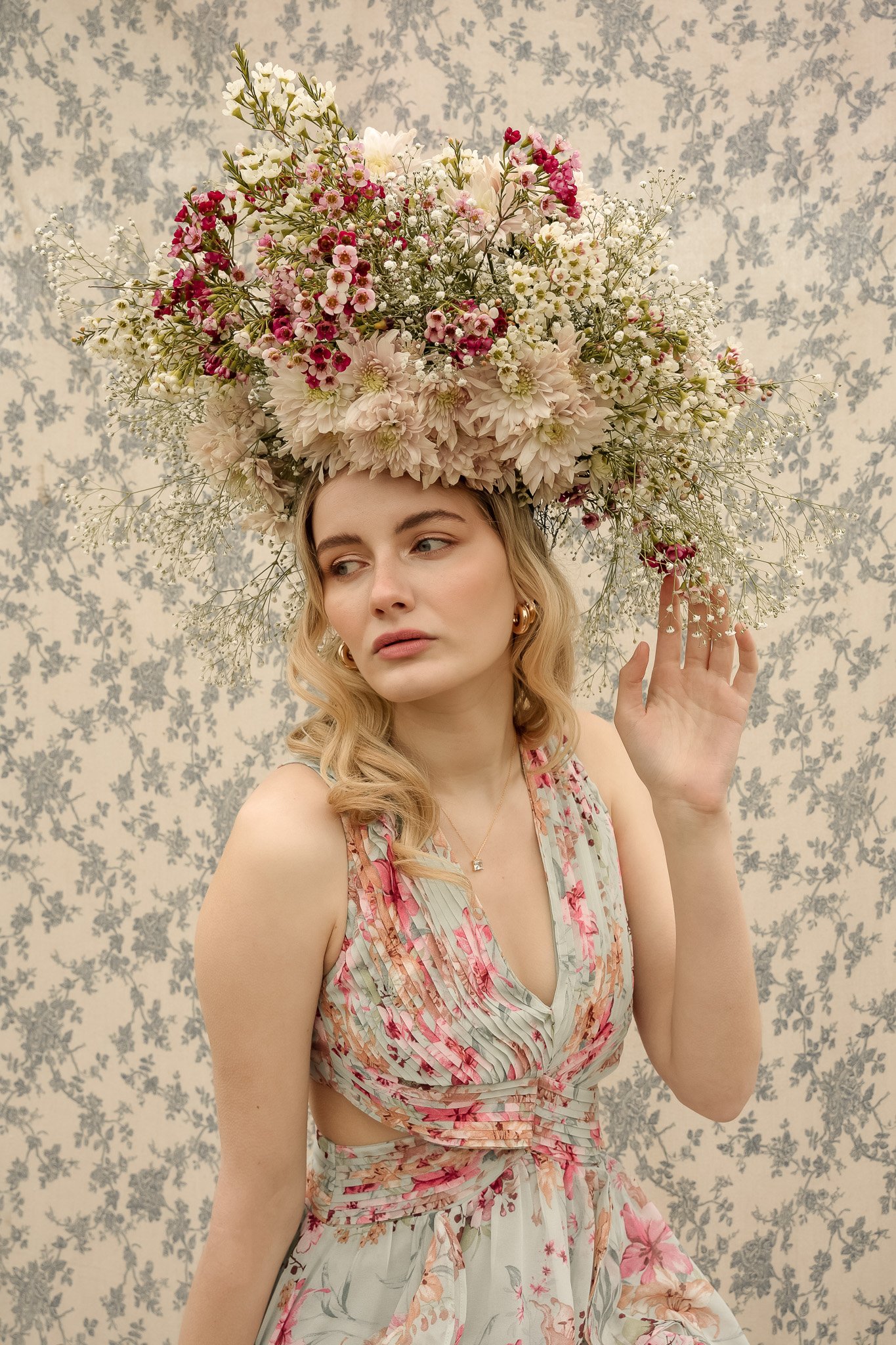Woman wearing a floral dress and elaborate flower crown, set against a floral wallpaper background.