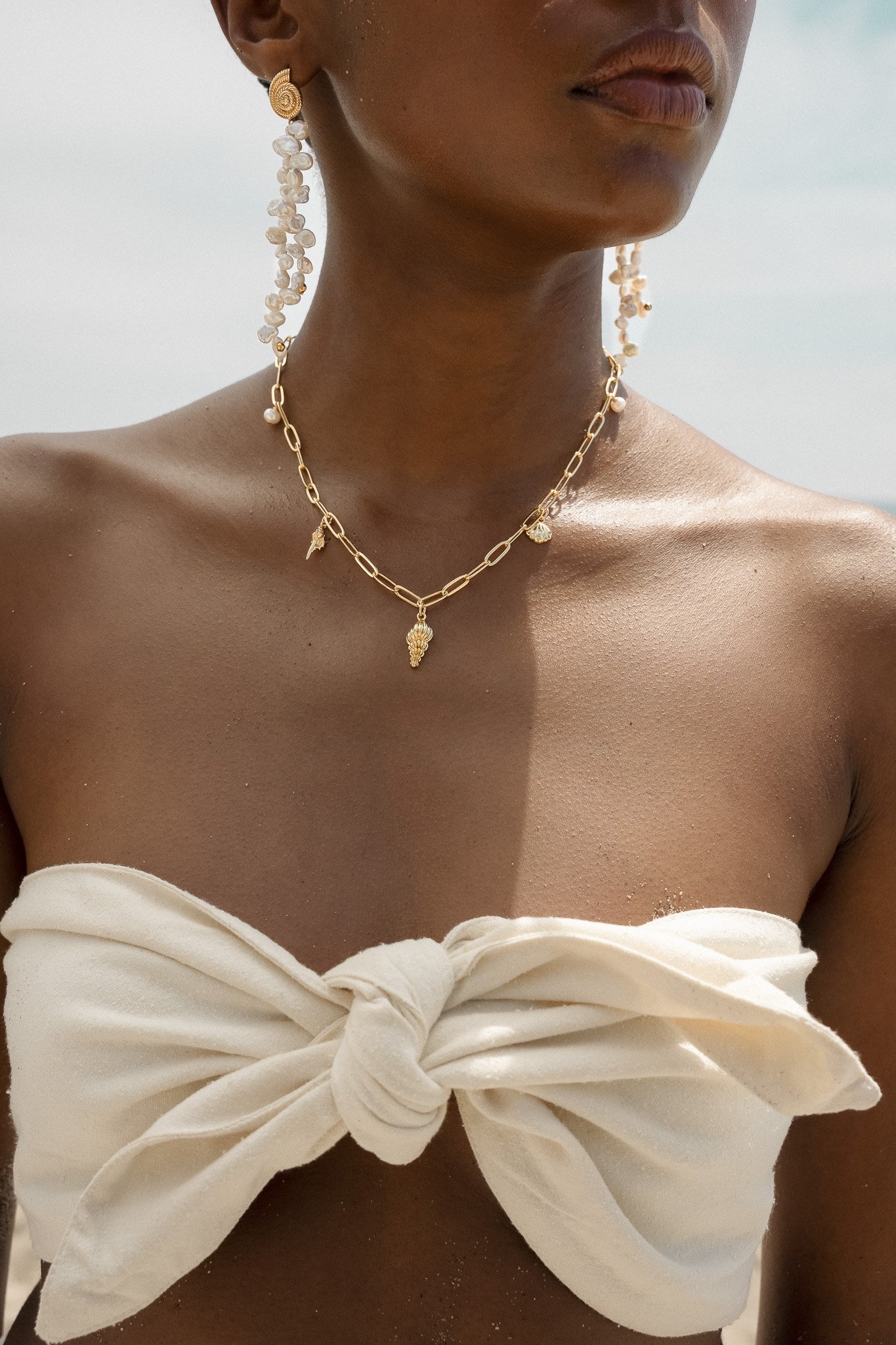 Close-up of a woman wearing a beige strapless top, a gold chain necklace with charms, and long beaded earrings.