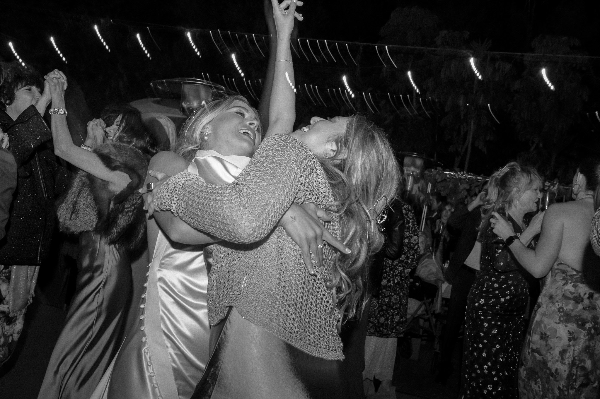 A black and white photo of people dancing at a festive event, with two women embracing and smiling in the foreground. Others are seen dancing and socializing in the background.