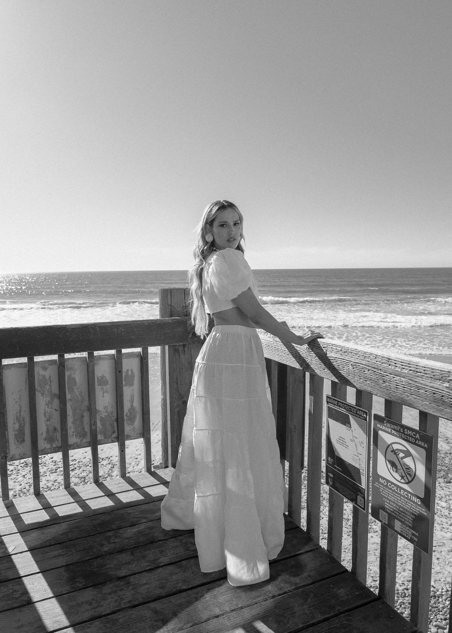 Woman in a long dress on a beach boardwalk, looking towards the camera, with the ocean in the background. Black and white photo.