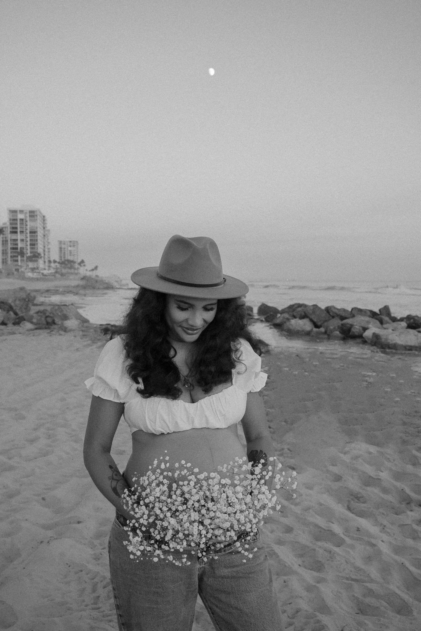 Pregnant woman wearing a hat and white top smiling, holding flowers on a sandy beach with moon and buildings in the background, black and white photo.