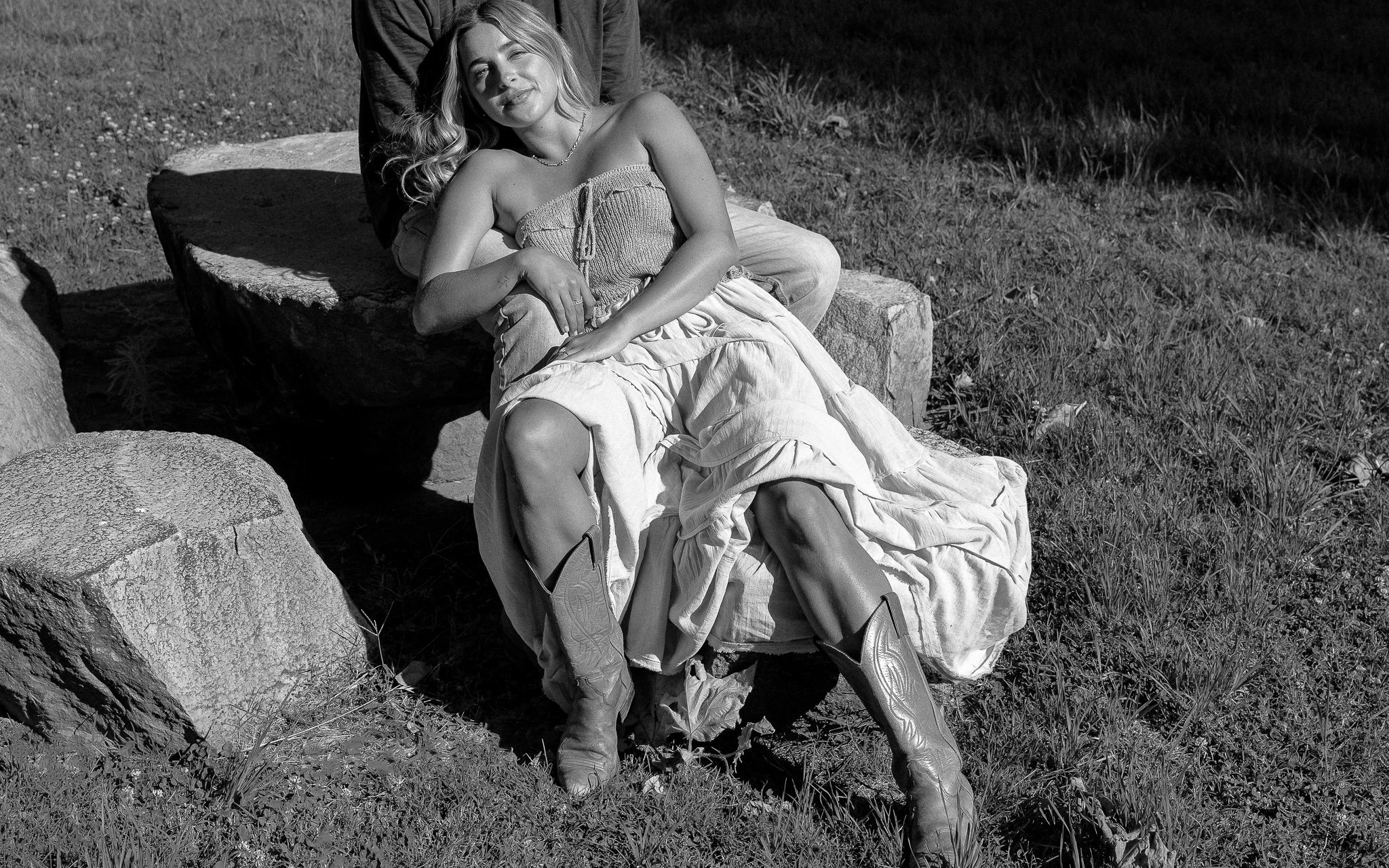 Woman in a dress and cowboy boots reclining on rocks outdoors in black and white.