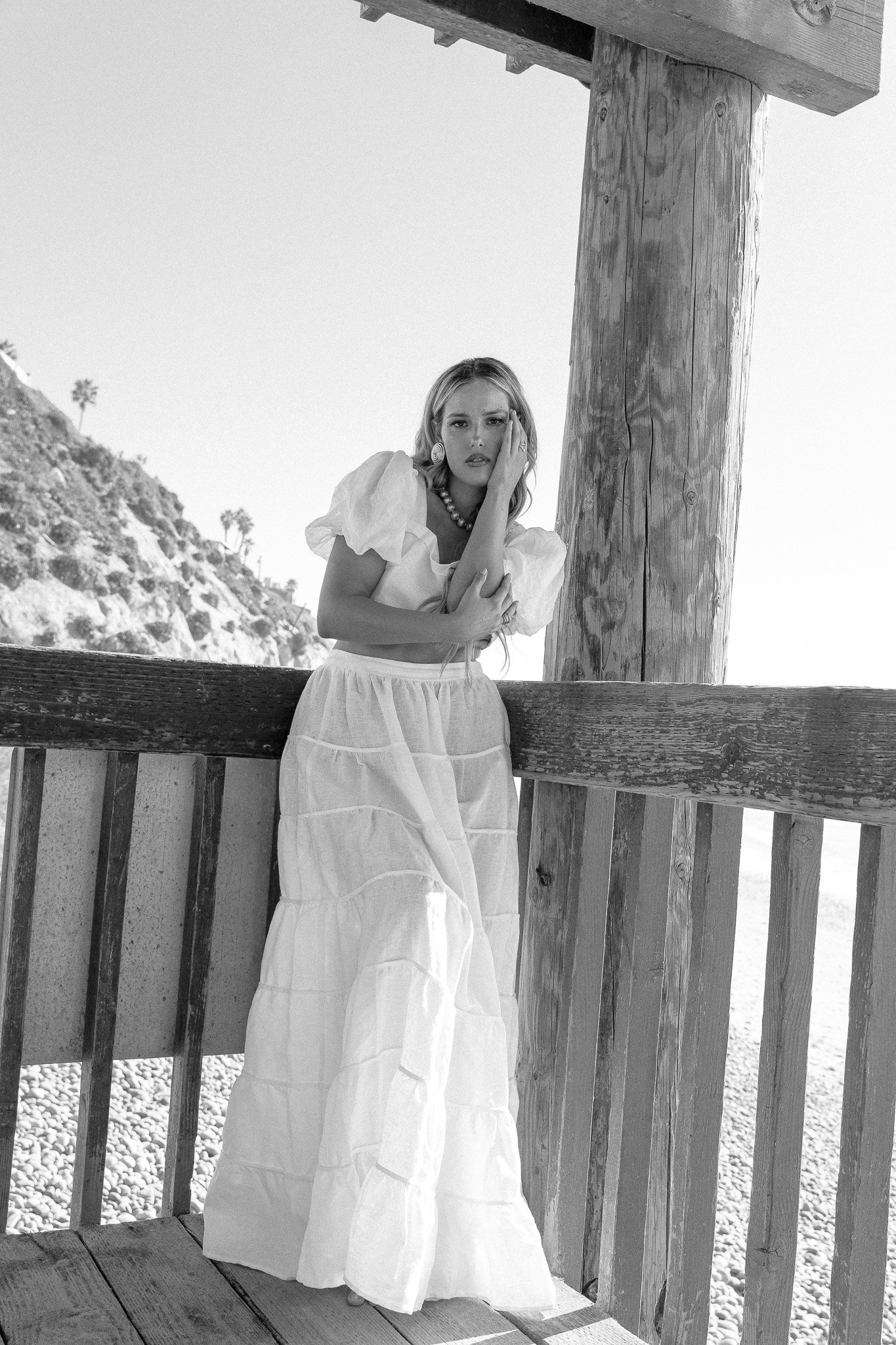 Black and white photo of a woman in a long white dress leaning against a wooden railing, with cliffs and palm trees in the background.