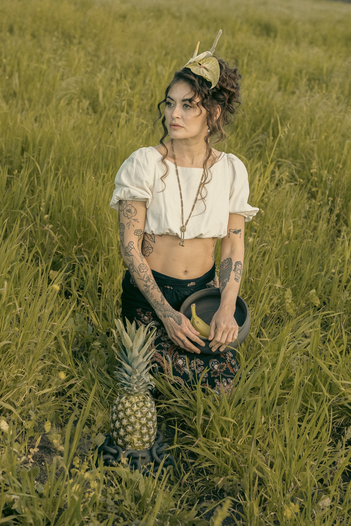 A woman with tattoos, wearing a white crop top and an ornate headpiece, sits in a grassy field holding a bowl with a banana. A pineapple is placed in front of her.
